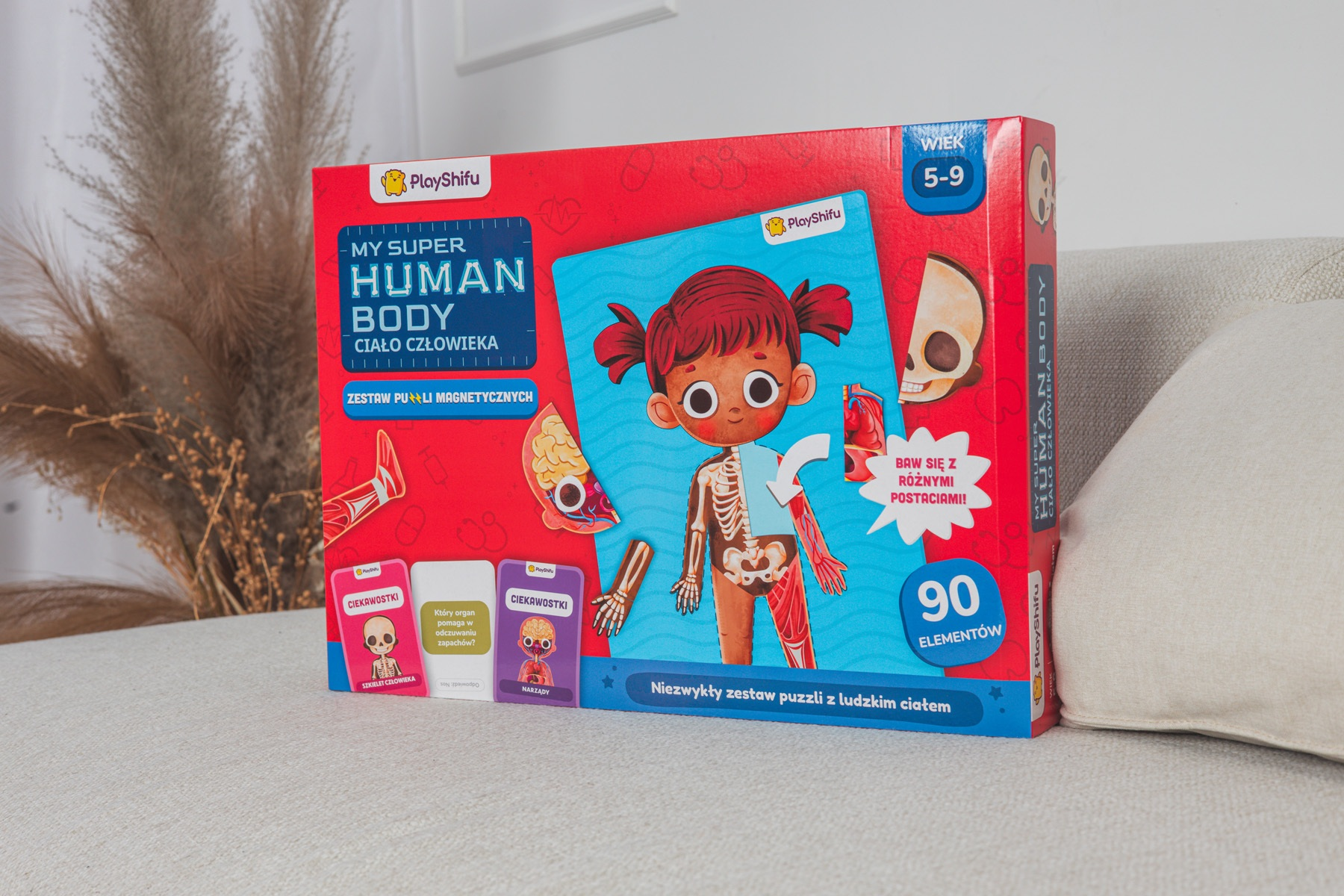 Zapakowana gra w puzzle o ciele człowieka. Zawiera ilustrację dziecka, podpisane części ciała i karty. Tło obejmuje sofę i suszoną roślinę.