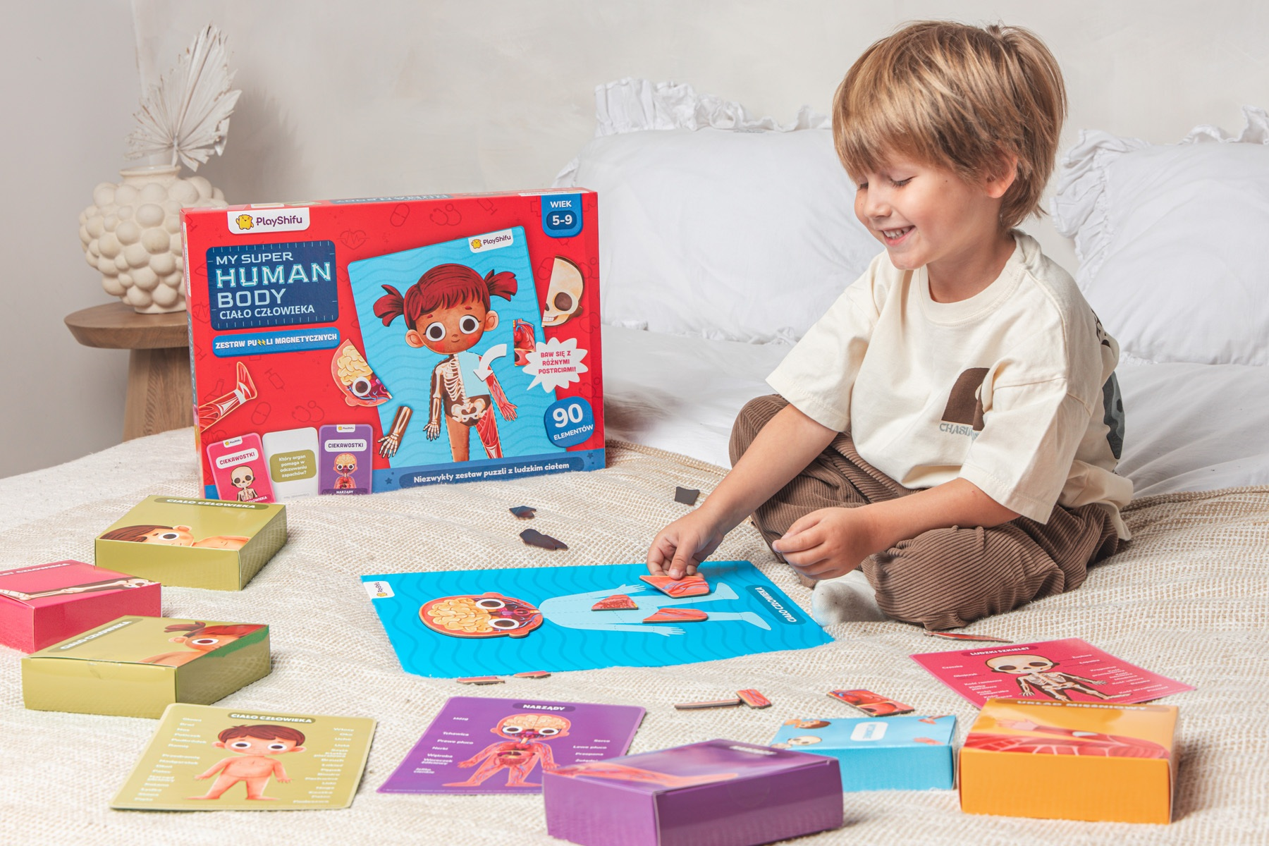 Uśmiechnięty chłopiec układa puzzle ludzkiego ciała. Scena obejmuje elementy układanki, pudełka i zestaw edukacyjny o ciele człowieka.