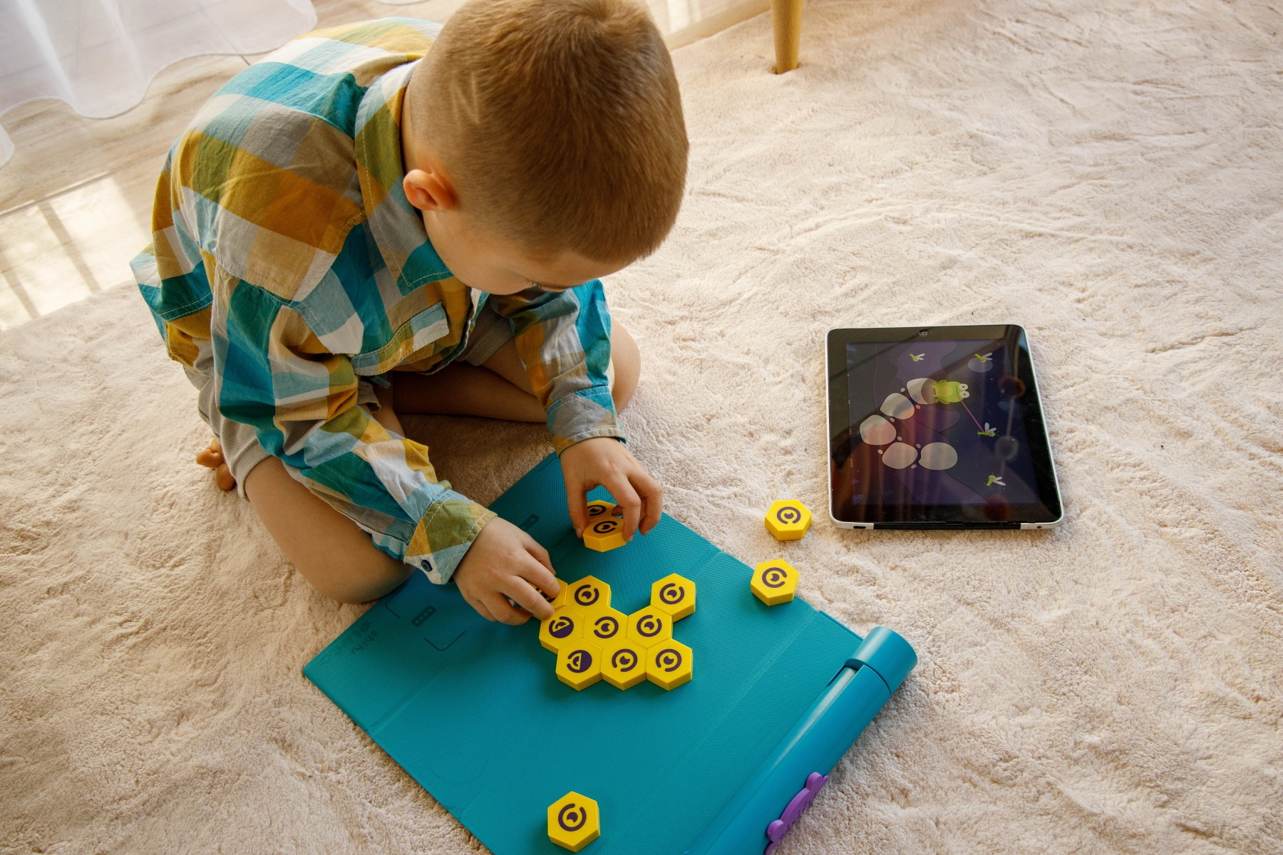 Dziecko bawi się żółtymi sześciokątnymi kafelkami na niebieskiej macie, w pobliżu tablet. Scena rozgrywa się na beżowym dywanie.
