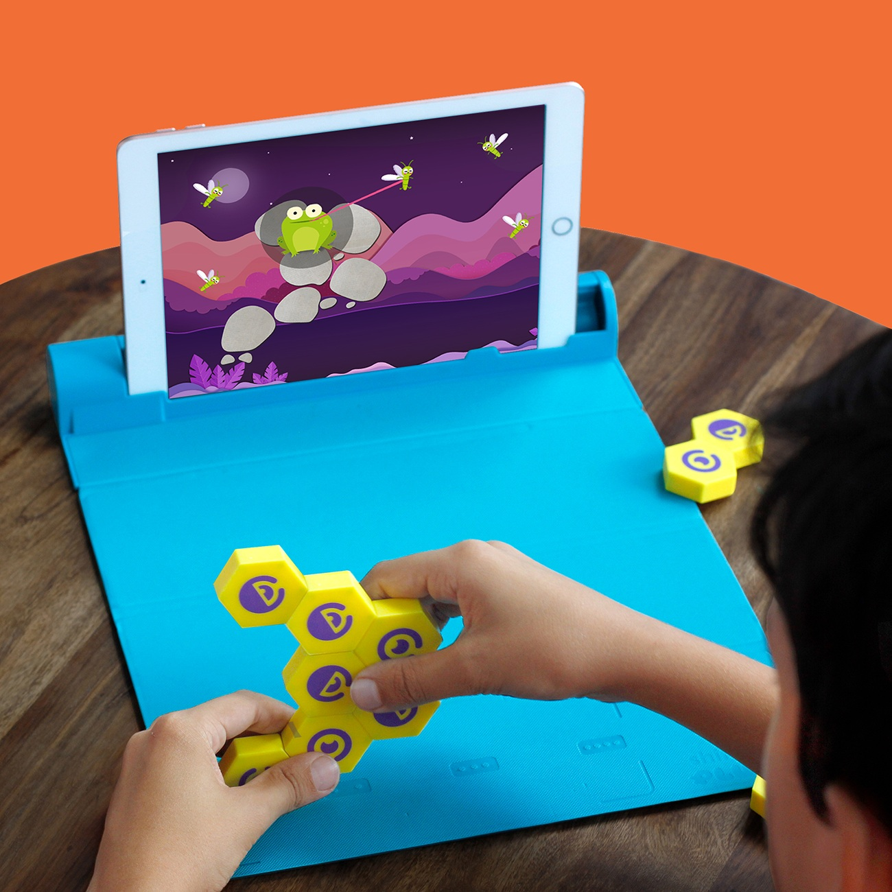 Dziecko trzyma żółte bloki sześciokątne z fioletowymi symbolami. Tablet pokazuje żabę łapiącą owady. Scena jest na niebieskiej powierzchni.
