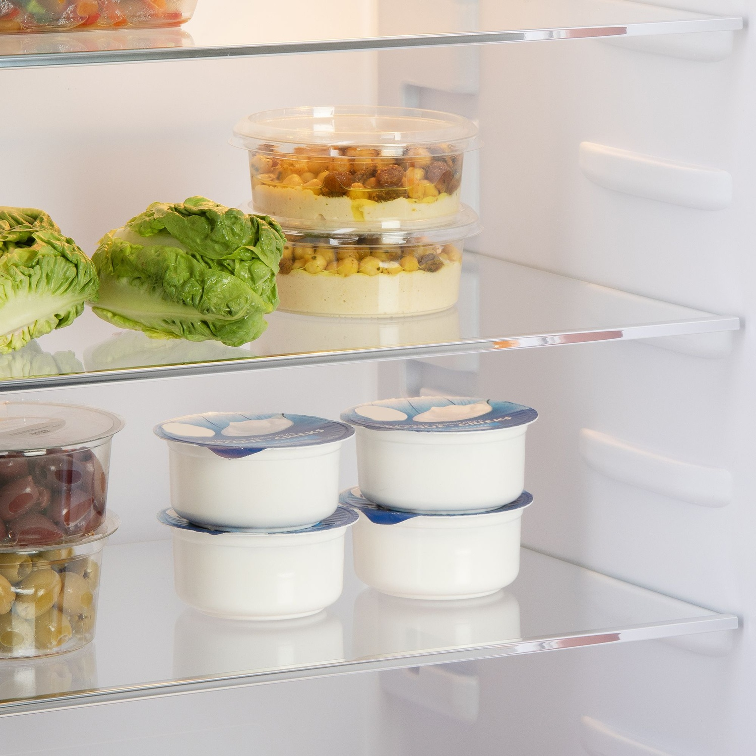 À l'intérieur d'un réfrigérateur : laitue, yaourt, houmous et olives. Les étagères sont en verre. Le réfrigérateur est blanc.