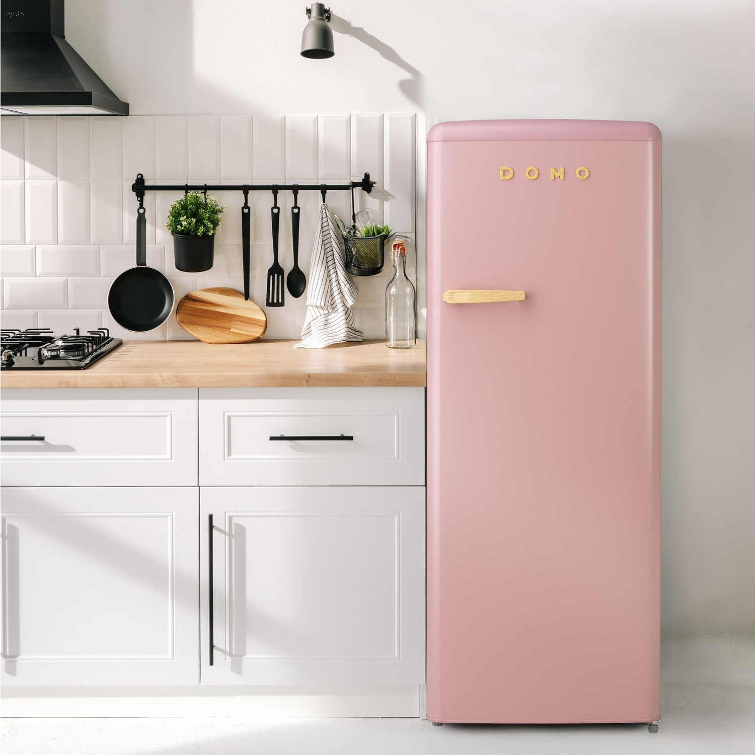 Un réfrigérateur rose rétro dans une cuisine avec des armoires blanches. Des ustensiles sont suspendus au-dessus d'un comptoir en bois, et une cuisinière à gaz à gauche.