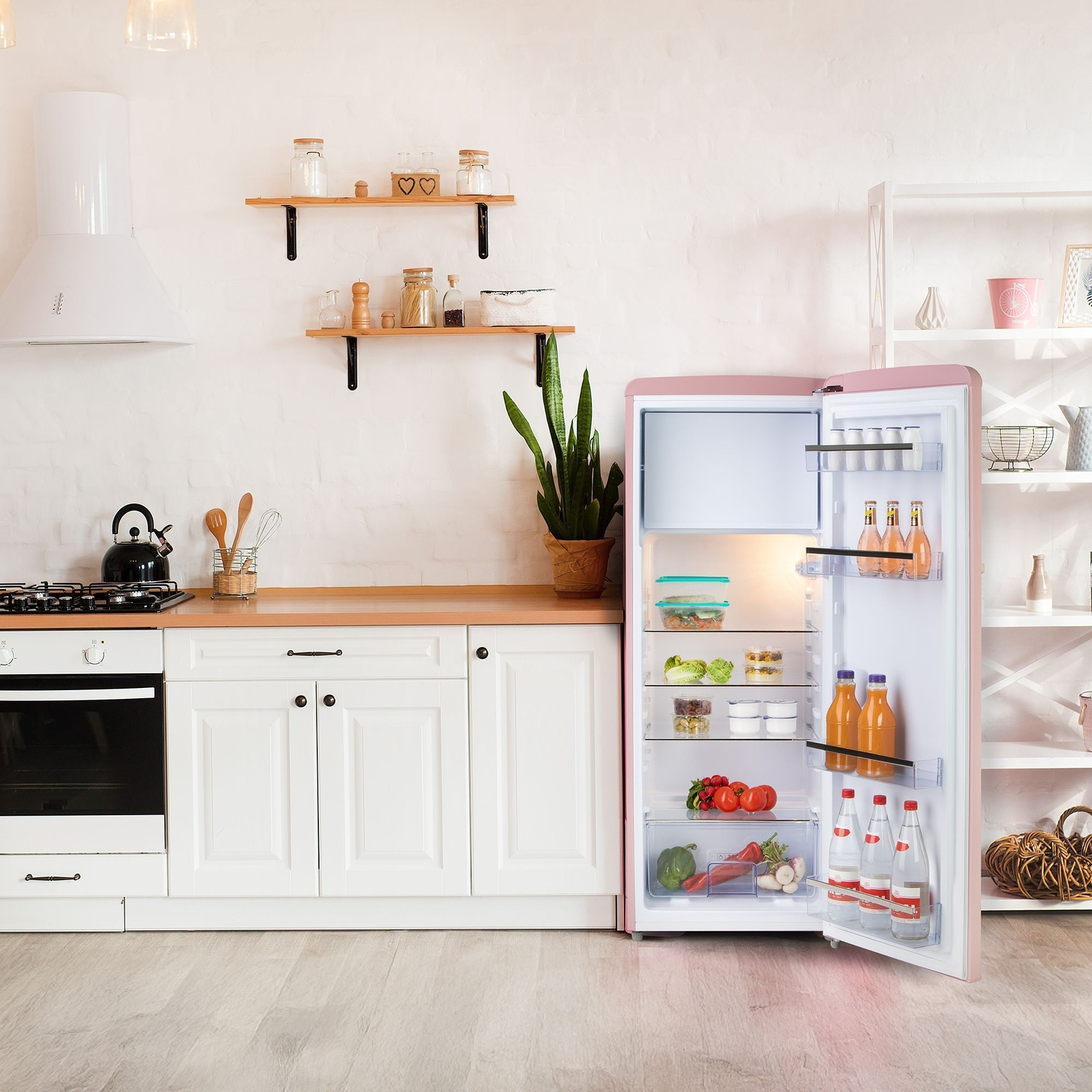 Cuisine avec réfrigérateur rose ouvert. À l'intérieur se trouvent des fruits, des légumes et des boissons. Armoires, cuisinière et étagères sont également visibles.