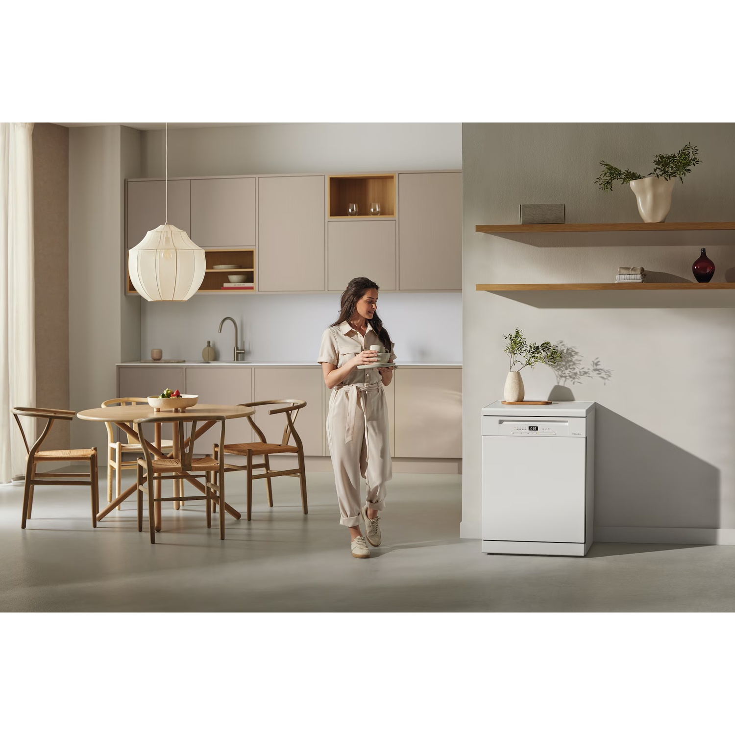 A woman in beige jumpsuit holds a cup in a modern kitchen. A round table with chairs and a white dishwasher are visible.