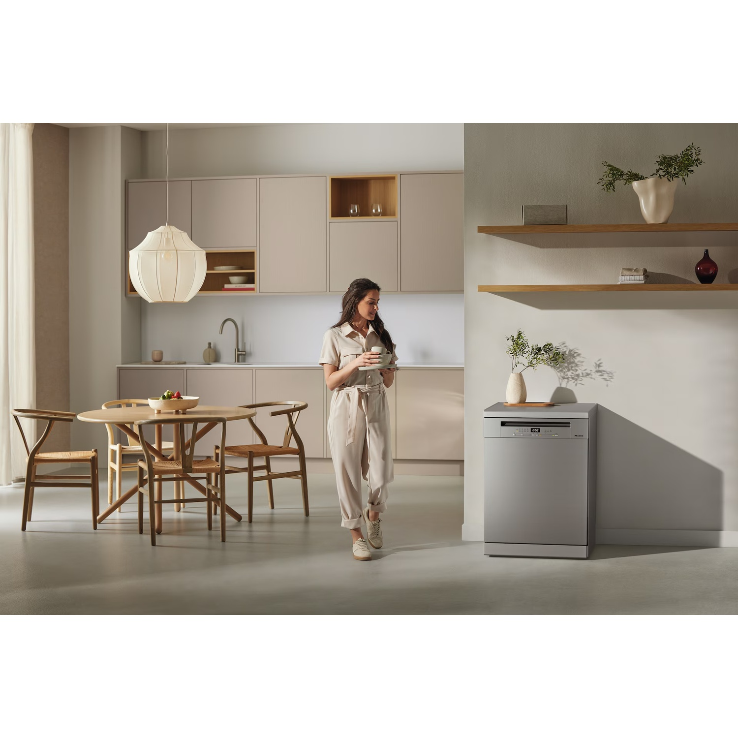 Woman in kitchen holding a cup. A round table with chairs is on the left. Dishwasher on the right.