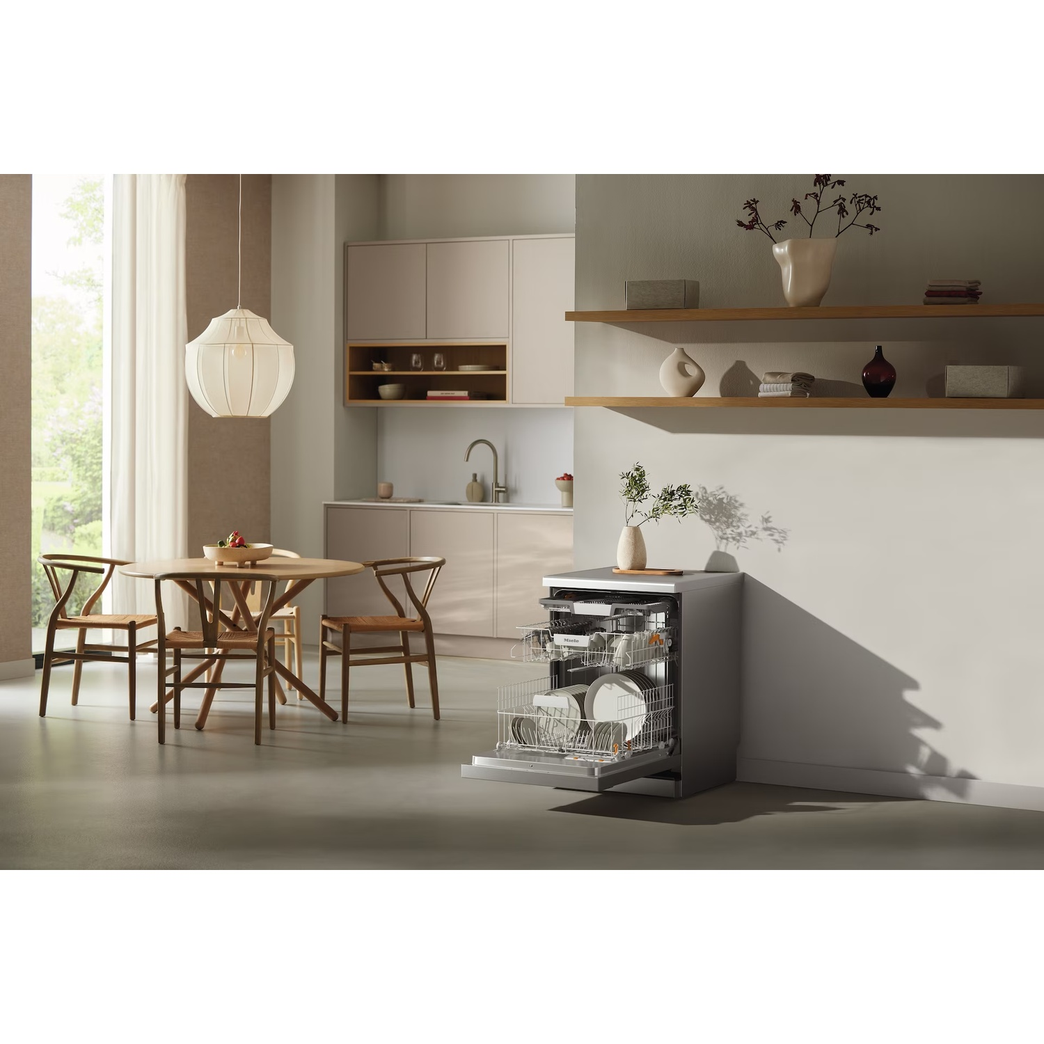 A modern kitchen with a dishwasher, table with chairs, and shelves with decorations. Light floods in from a window.