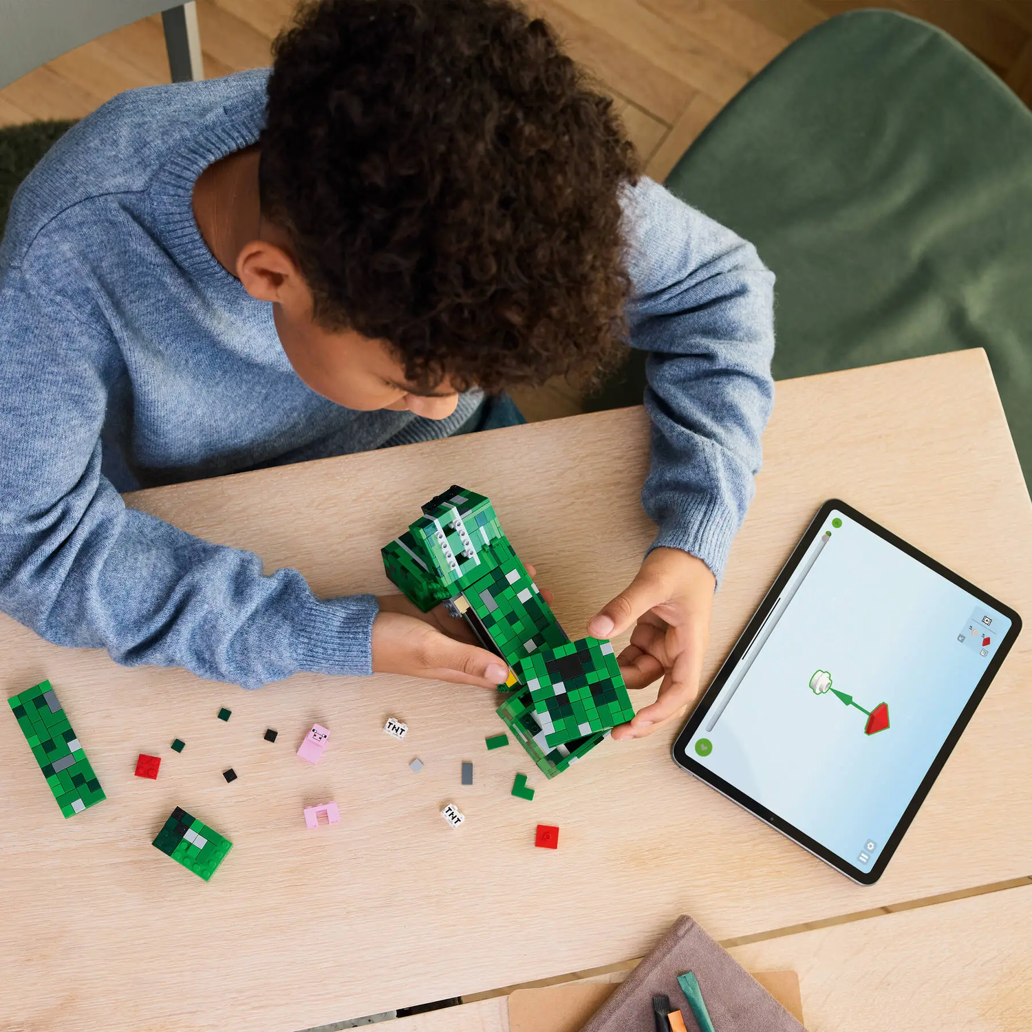 Een persoon bouwt een groen Minecraft-figuur met blokken, gebruikt een tablet voor instructies, op een houten tafel. Kleine blokken verspreid.