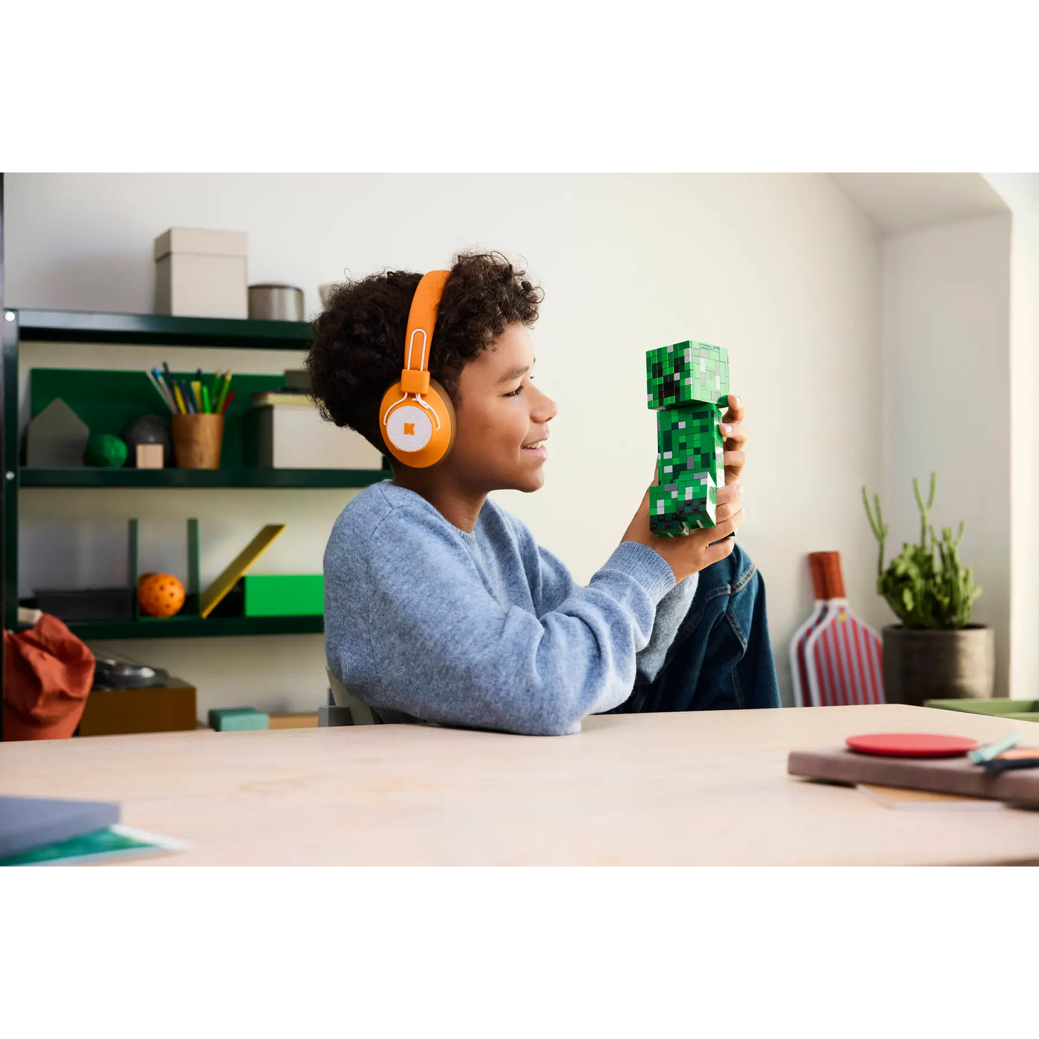 Een jongen met een oranje koptelefoon houdt een Minecraft Creeper-figuur vast. Hij glimlacht terwijl hij aan een tafel zit. Op de achtergrond staan planken.