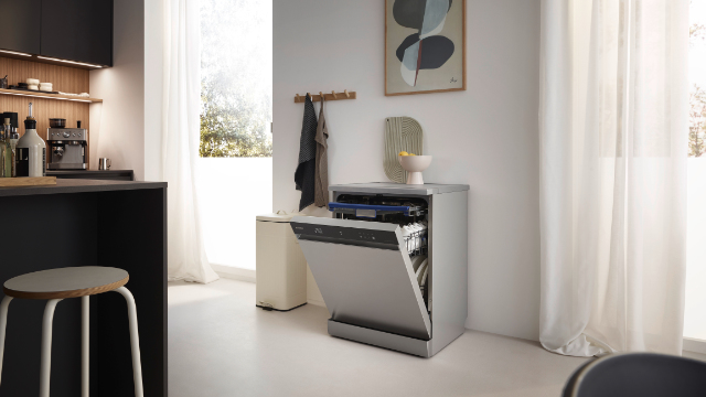 Otwarta zmywarka we współczesnej kuchni. W tle czarny blat ze stołkiem i okno z białymi zasłonami.