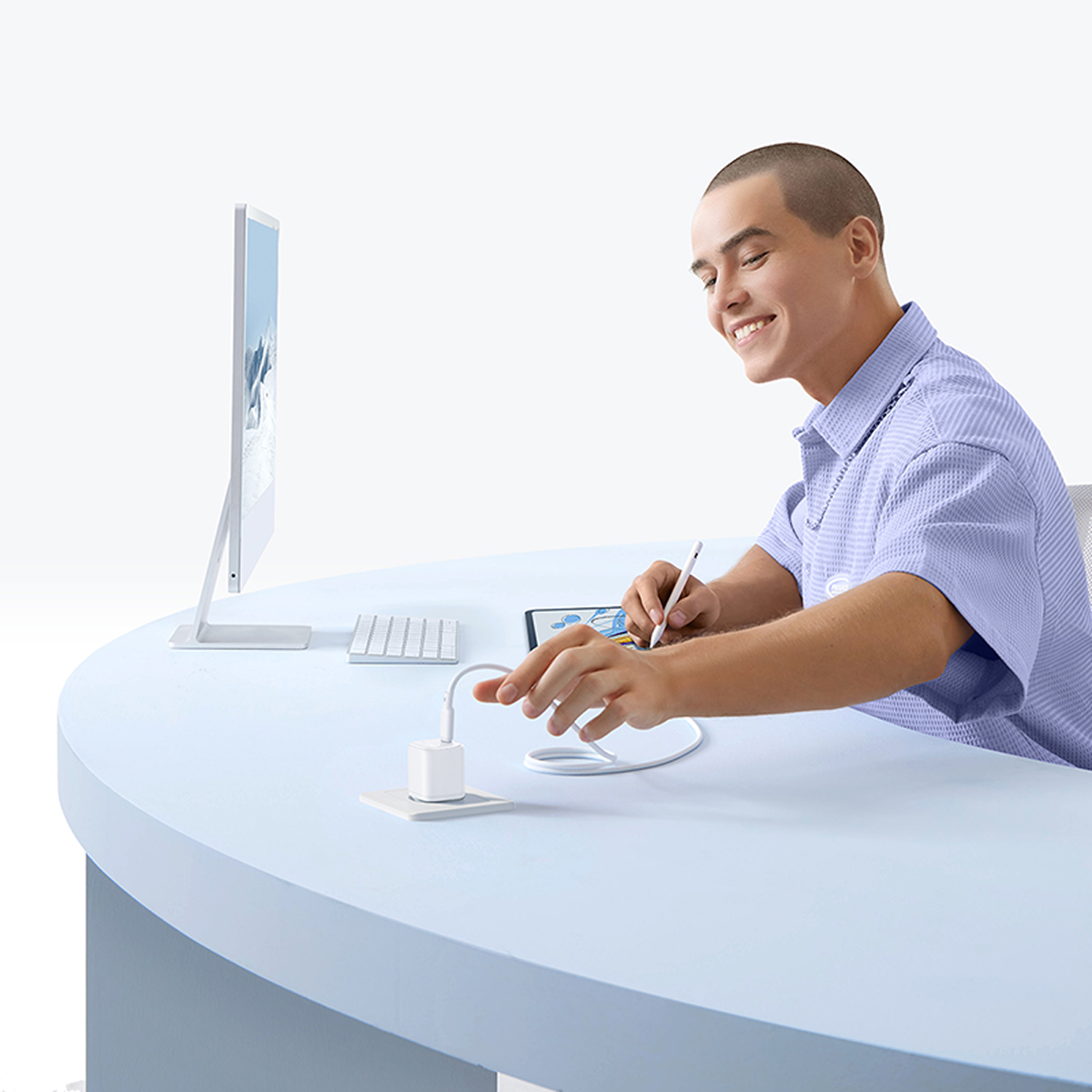 Un hombre sonríe, usando una tableta con un lápiz óptico. Monitor, teclado y cargador sobre una mesa azul. Fondo blanco.