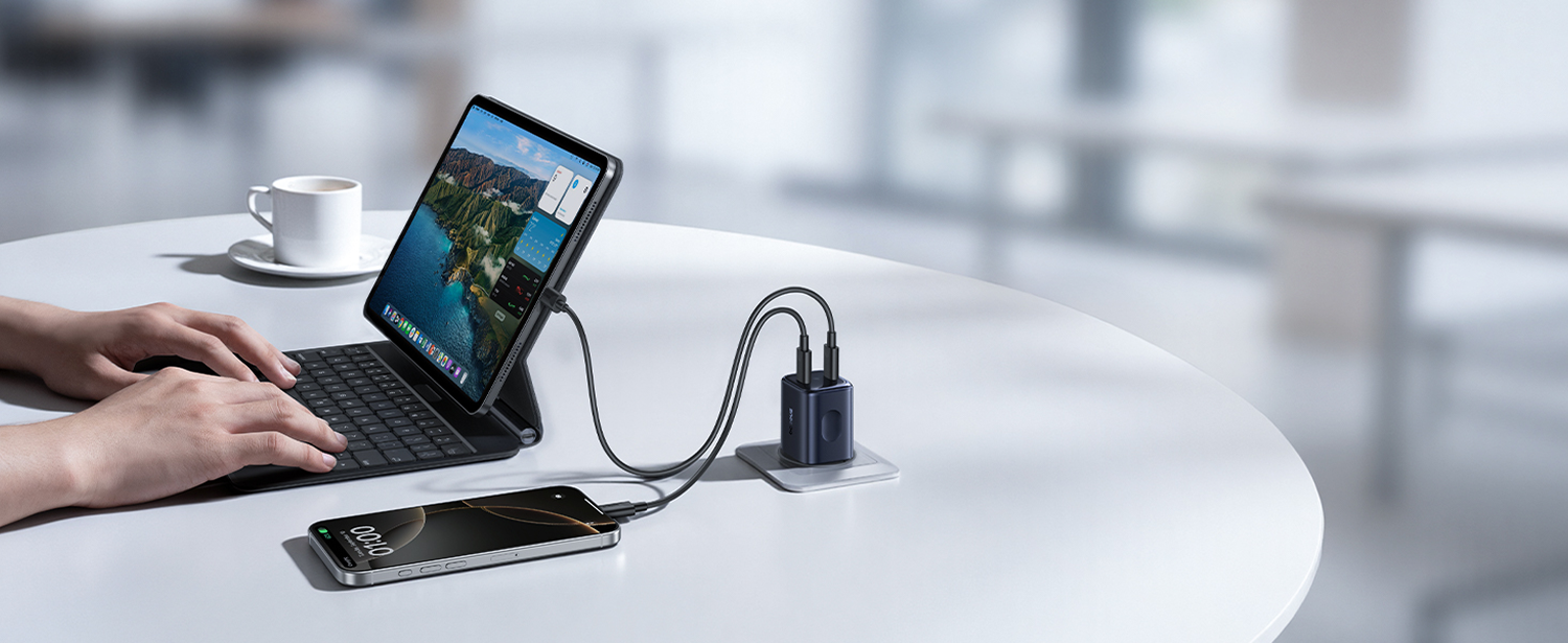 Una tableta y un teléfono se cargan en una mesa blanca. Una persona usa la tableta con un teclado. Una taza está cerca.