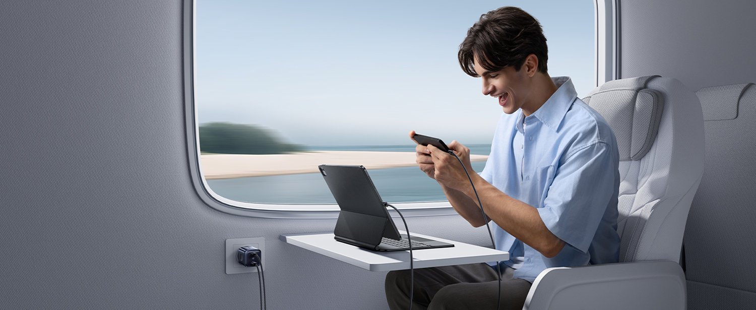 Un hombre en un tren juega con un teléfono y una tableta. Se ve agua y cielo por la ventana.