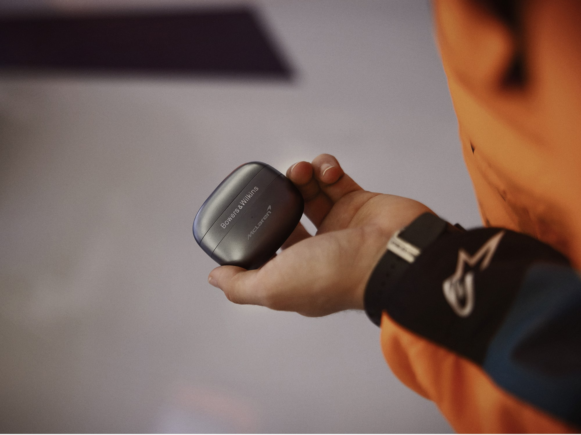 Una mano sostiene un estuche de auriculares Bowers & Wilkins McLaren gris. La persona lleva un traje de carreras naranja y una pulsera negra.