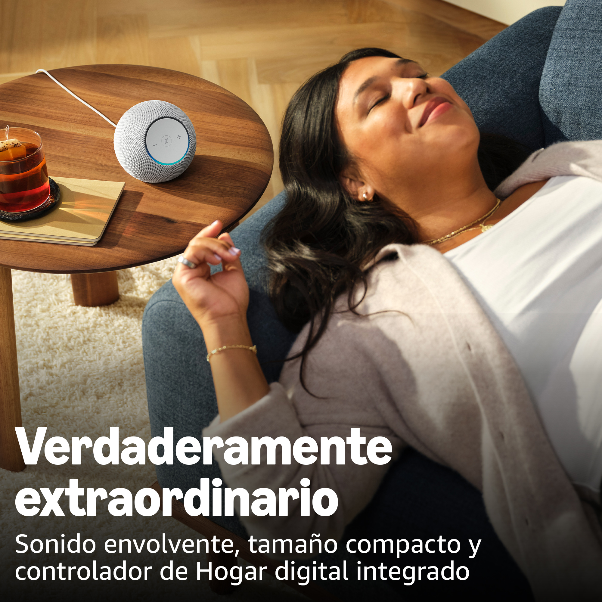 Mujer relajándose en un sofá azul, un altavoz inteligente sobre una mesa de madera, con una taza de té. Texto: 'Verdaderamente extraordinario'.