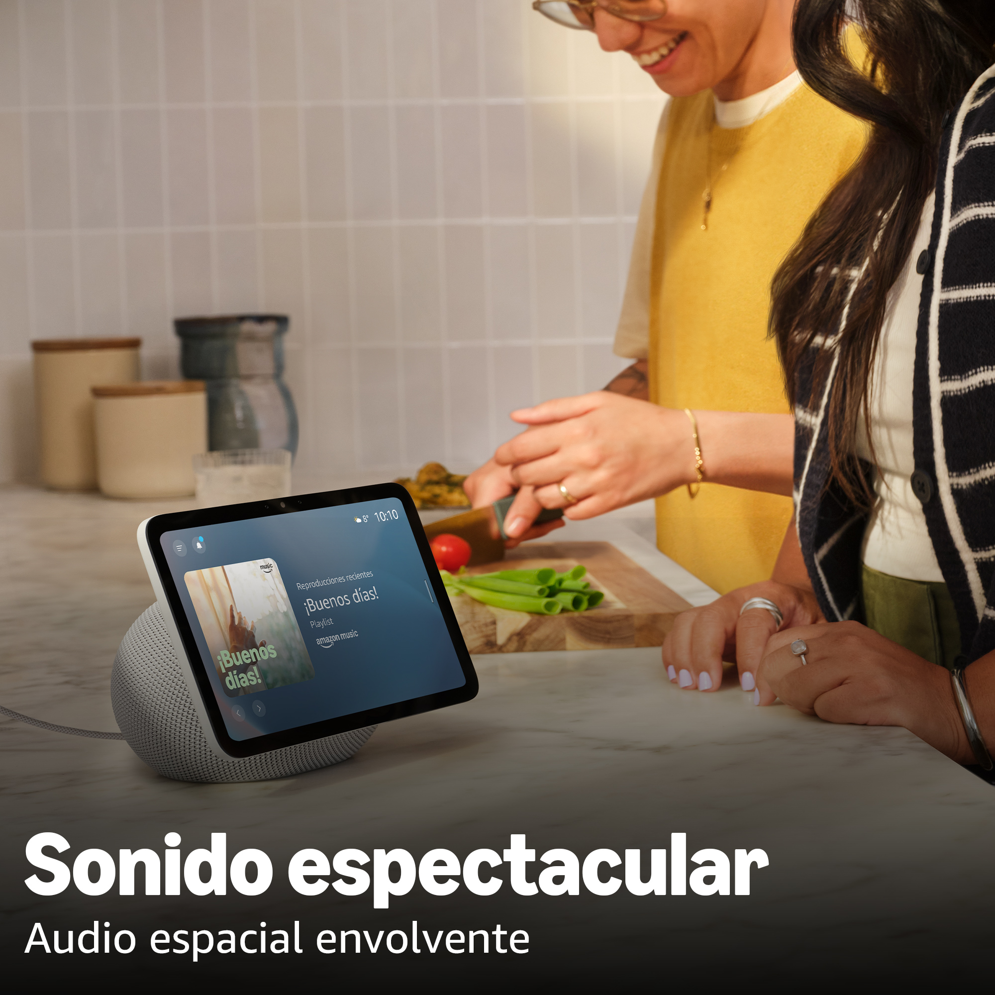 Un altavoz inteligente con pantalla muestra una lista de reproducción. Dos personas cortan verduras en el fondo.