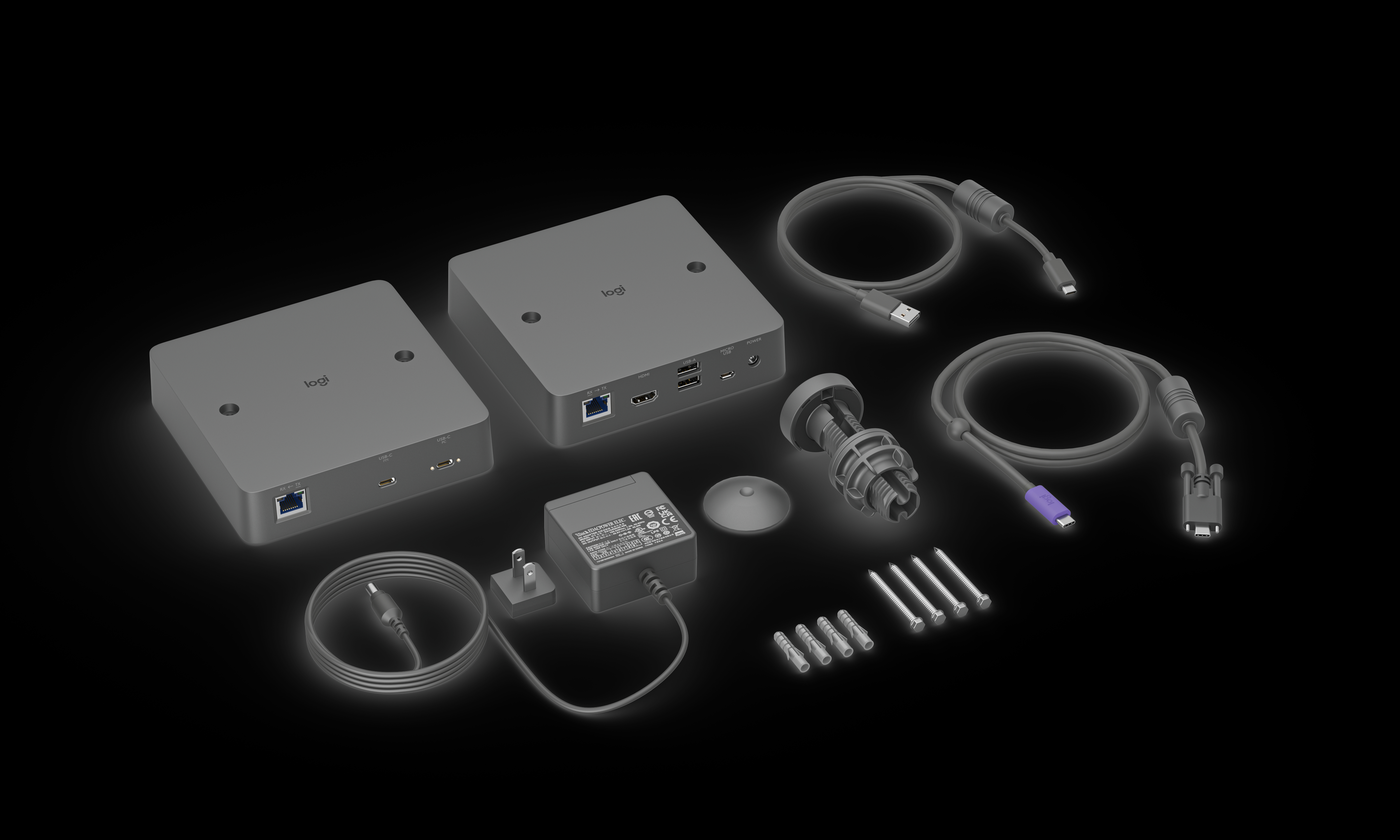 Das Bild zeigt zwei graue Logitech-Geräte mit verschiedenen Kabeln, einem Netzteil, Schrauben und Befestigungsmaterial vor schwarzem Hintergrund.