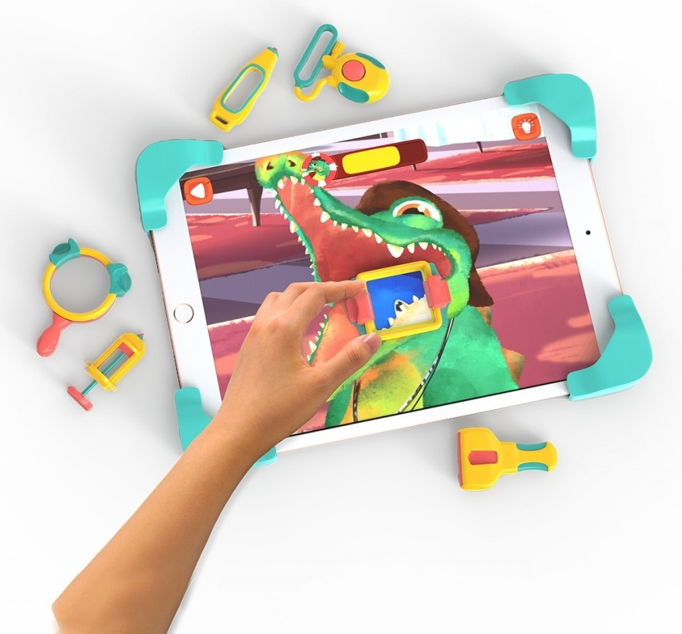 Ręka wchodzi w interakcję z tabletem pokazującym kreskówkowego aligatora. Zabawki otaczają tablet: stetoskop, lupa i strzykawka.