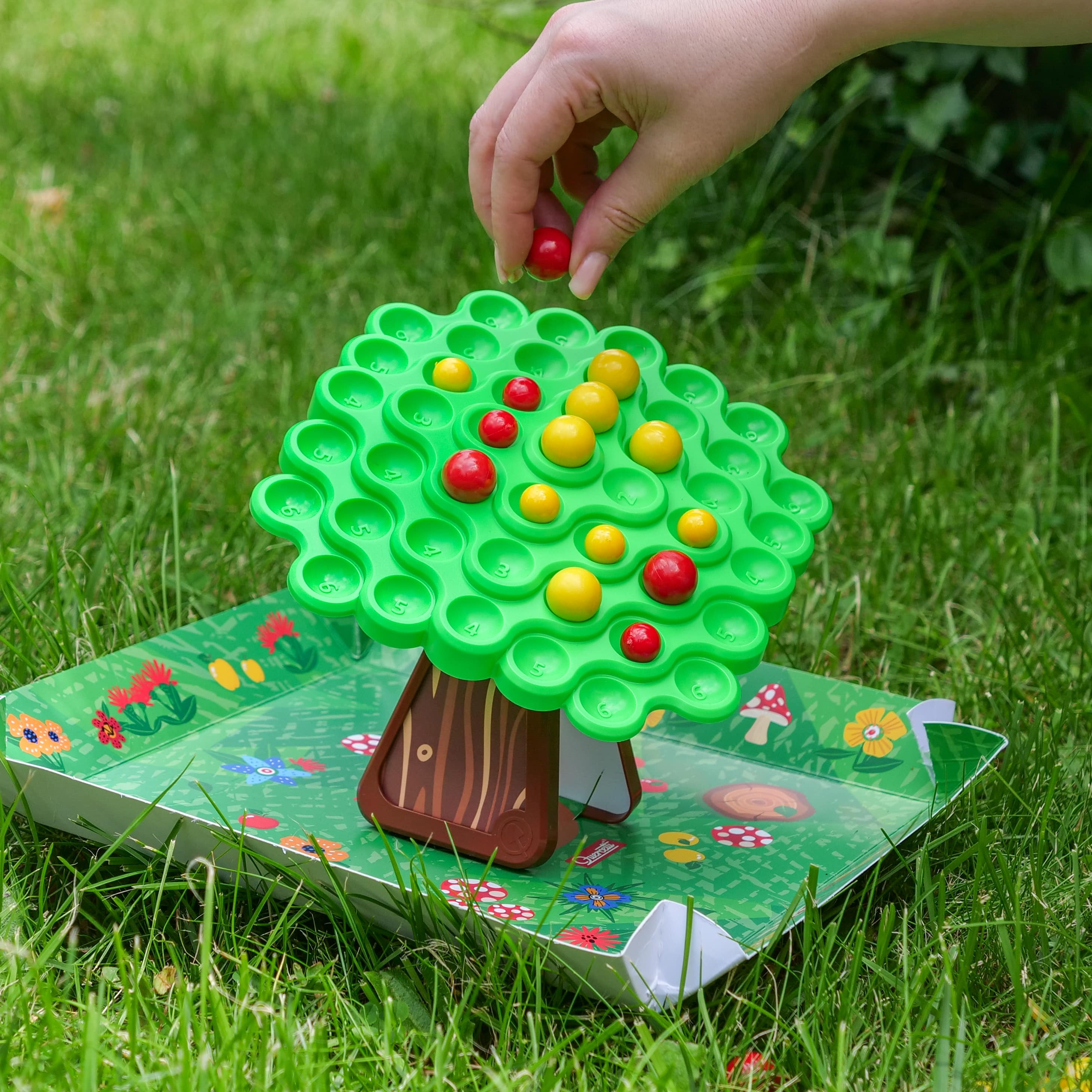 Ręka umieszcza czerwoną kulkę na zielonej grze w kształcie drzewa. Gra stoi na zielonej macie na trawie.