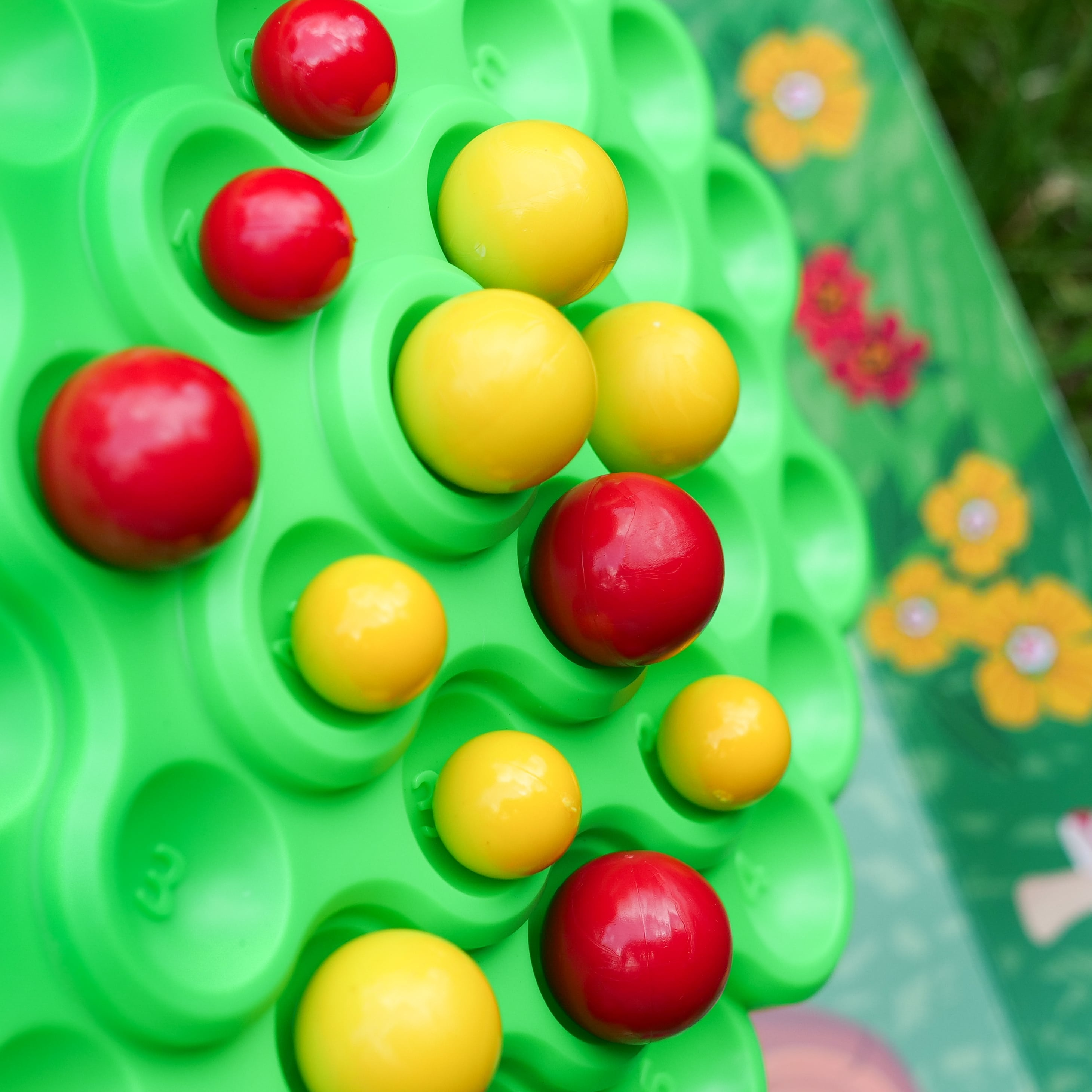 Zielona plansza z czerwonymi i żółtymi kulkami. Niektóre kulki są w otworach. W tle rozmyte kwiaty.