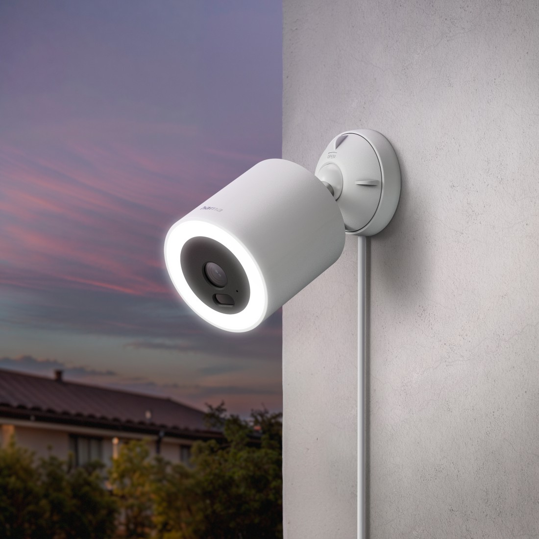 Eine weiße Überwachungskamera mit leuchtendem Ring ist an einer Wand montiert. Im Hintergrund sind Sonnenuntergang und ein Haus zu sehen.