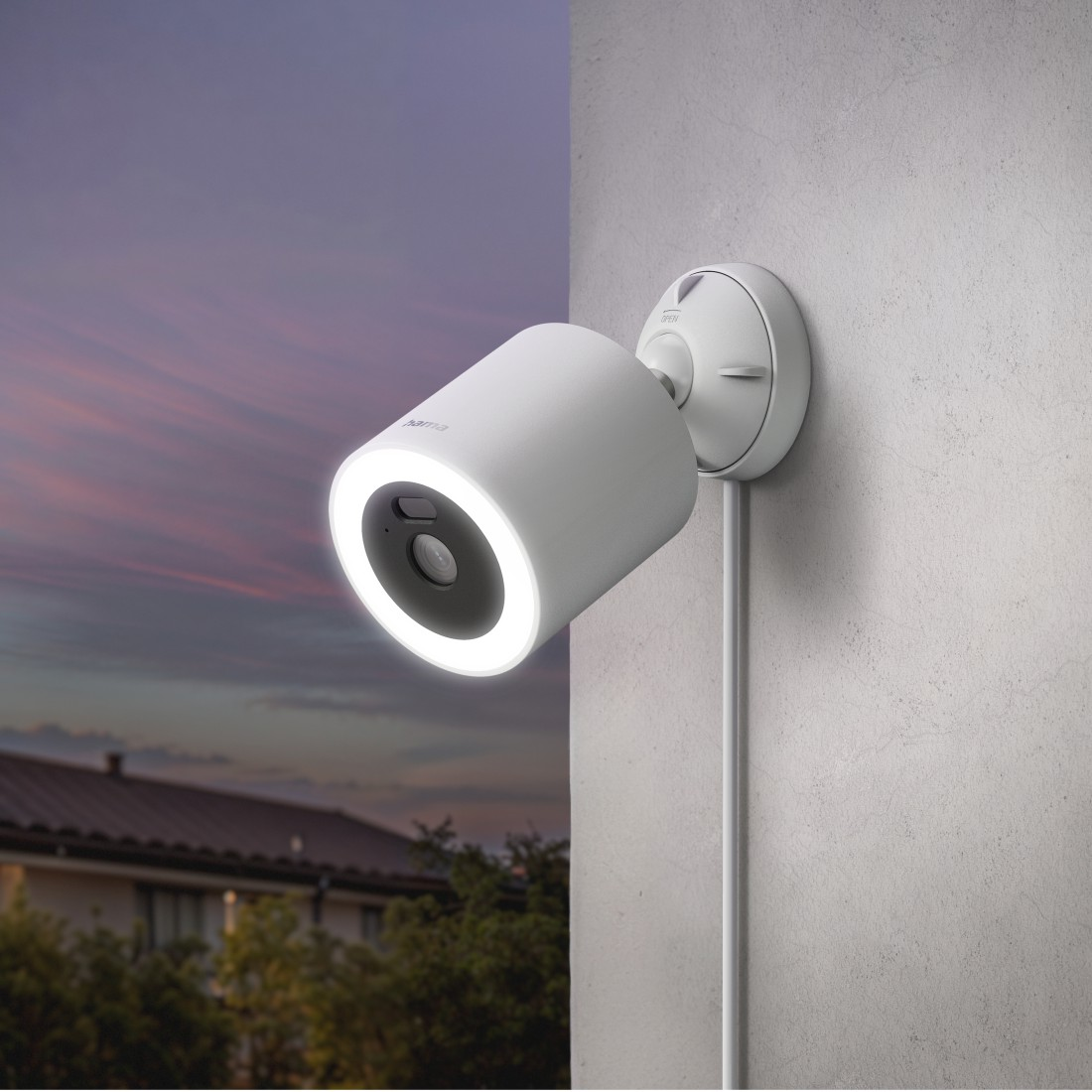 Eine weiße Überwachungskamera mit beleuchtetem Ring an einer Wand. Abendhimmel im Hintergrund mit einer Haussilhouette.