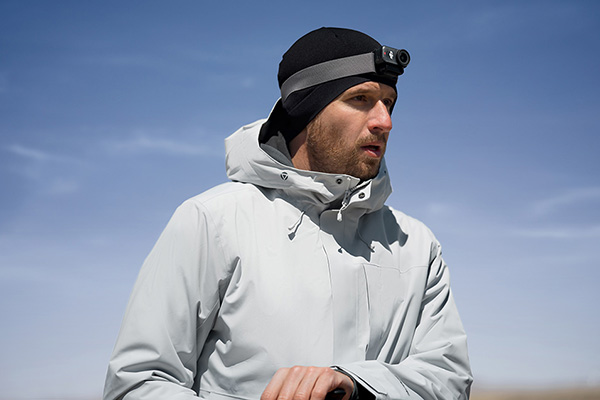 Mężczyzna w czarnej czapce z czołówką i jasno szarej kurtce stoi na zewnątrz na tle błękitnego nieba.