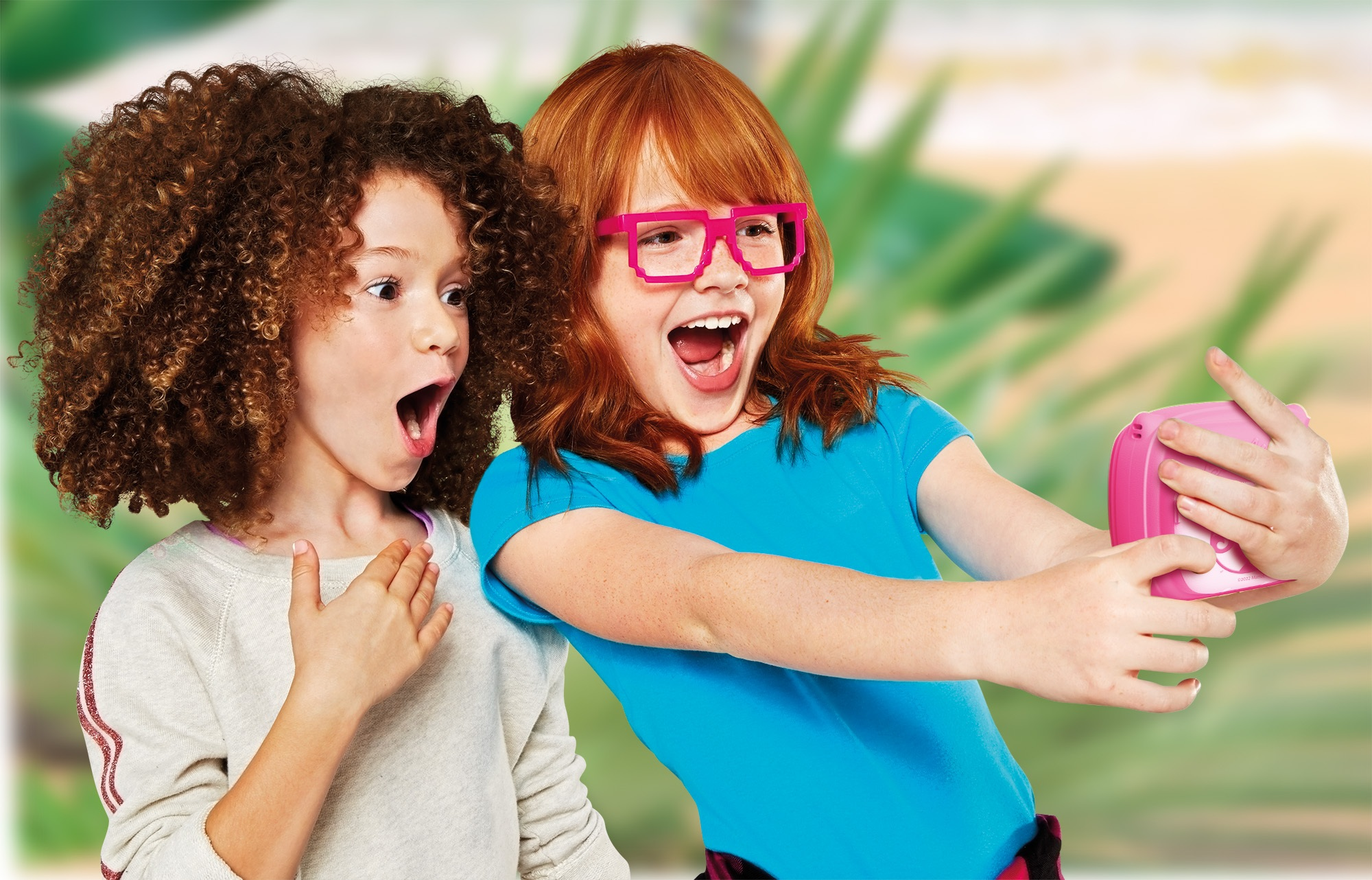 Dwie dziewczyny są podekscytowane. Jedna w okularach trzyma różowy aparat. Druga ma kręcone włosy. Obie mają otwarte usta.