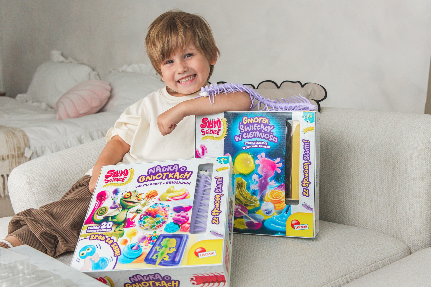 Uśmiechnięty chłopiec siedzi z dwoma zestawami naukowymi, jednym z fioletową zabawką stonogą. Tło obejmuje łóżko i sofę.