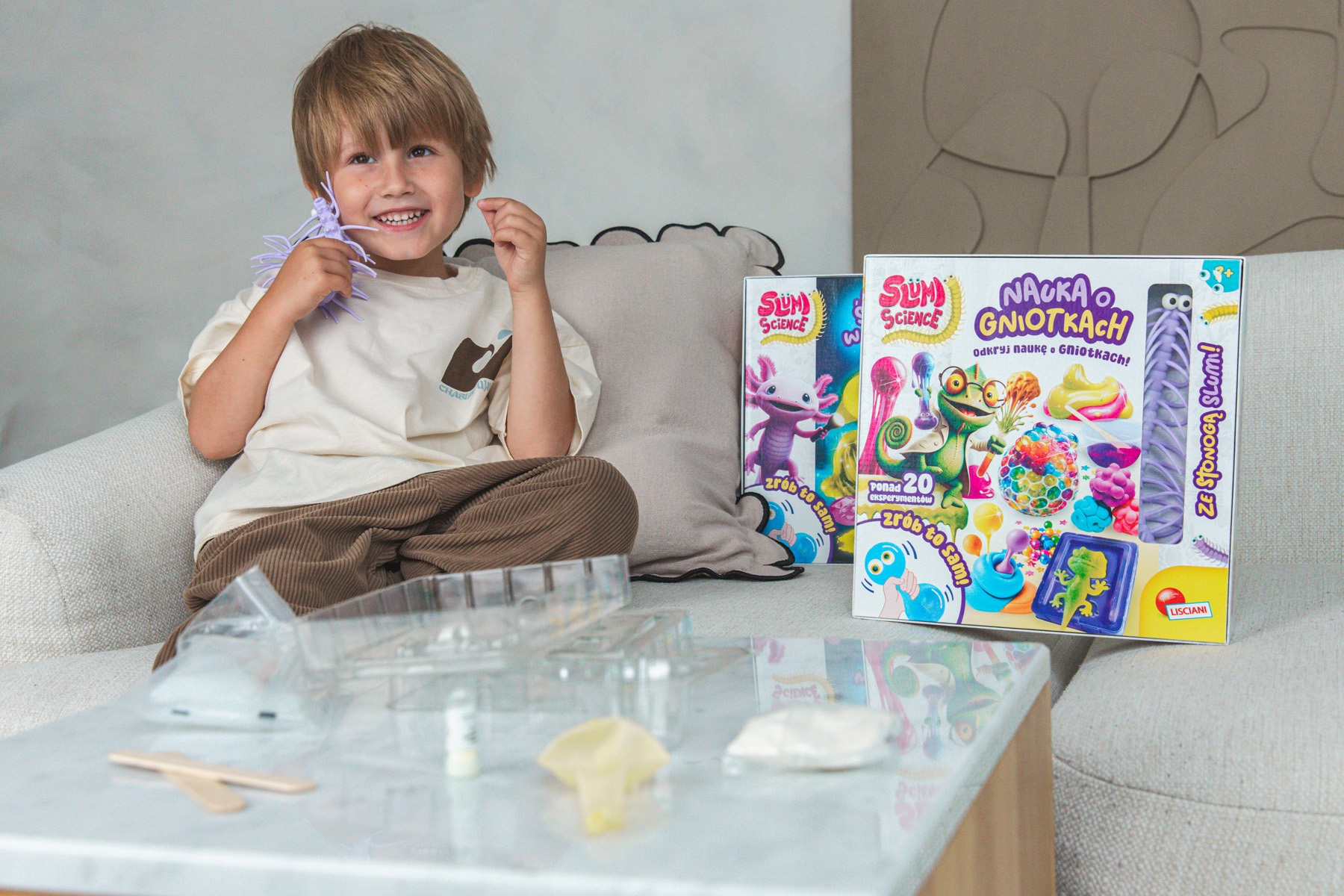 Uśmiechnięty chłopiec trzyma fioletową zabawkę. Za nim pudełka z eksperymentami naukowymi. Na stole materiały.