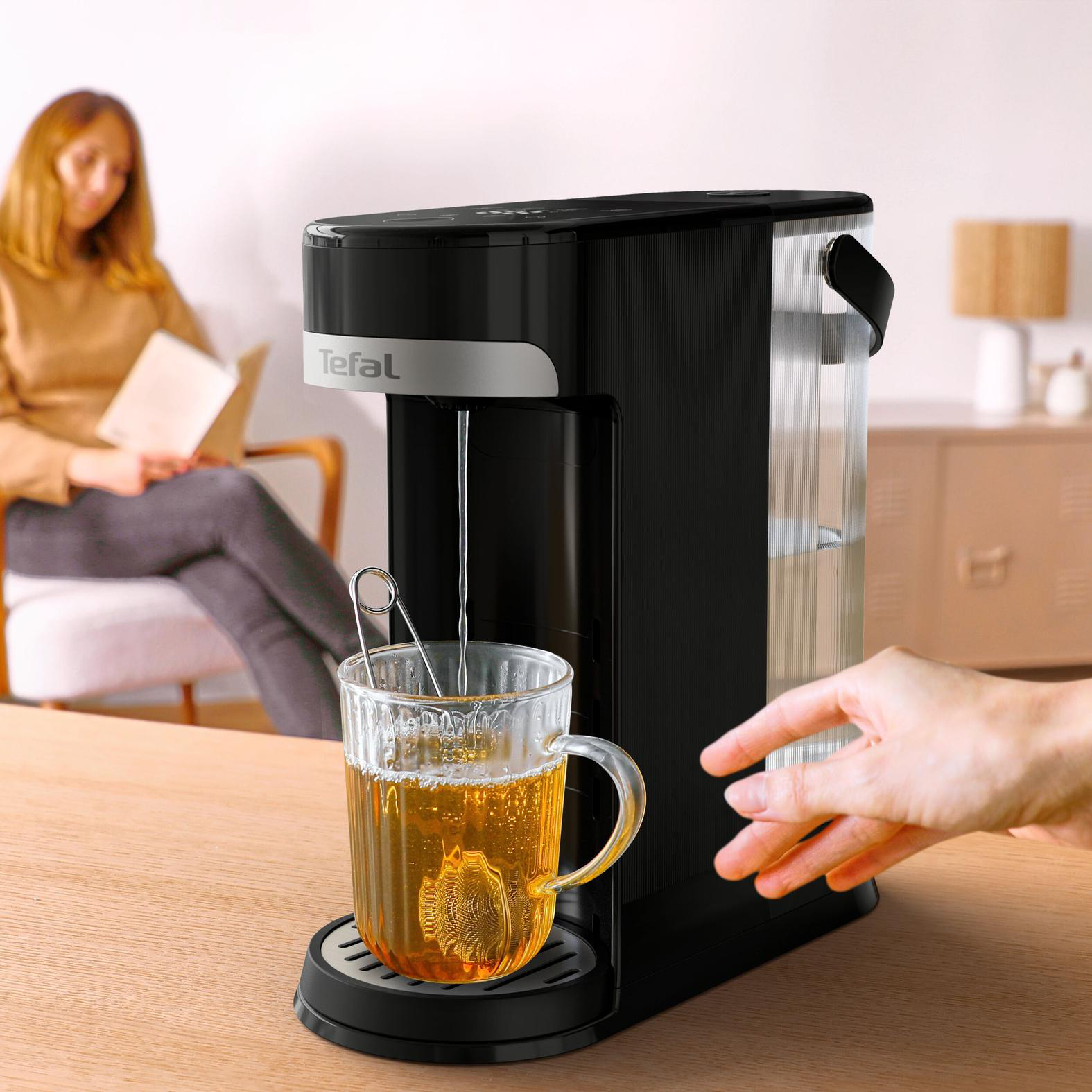 Ein schwarzer und silberner Tefal-Teekocher mit einem Glas Tee. Im Hintergrund liest eine Person, und die Hand einer anderen Person greift.