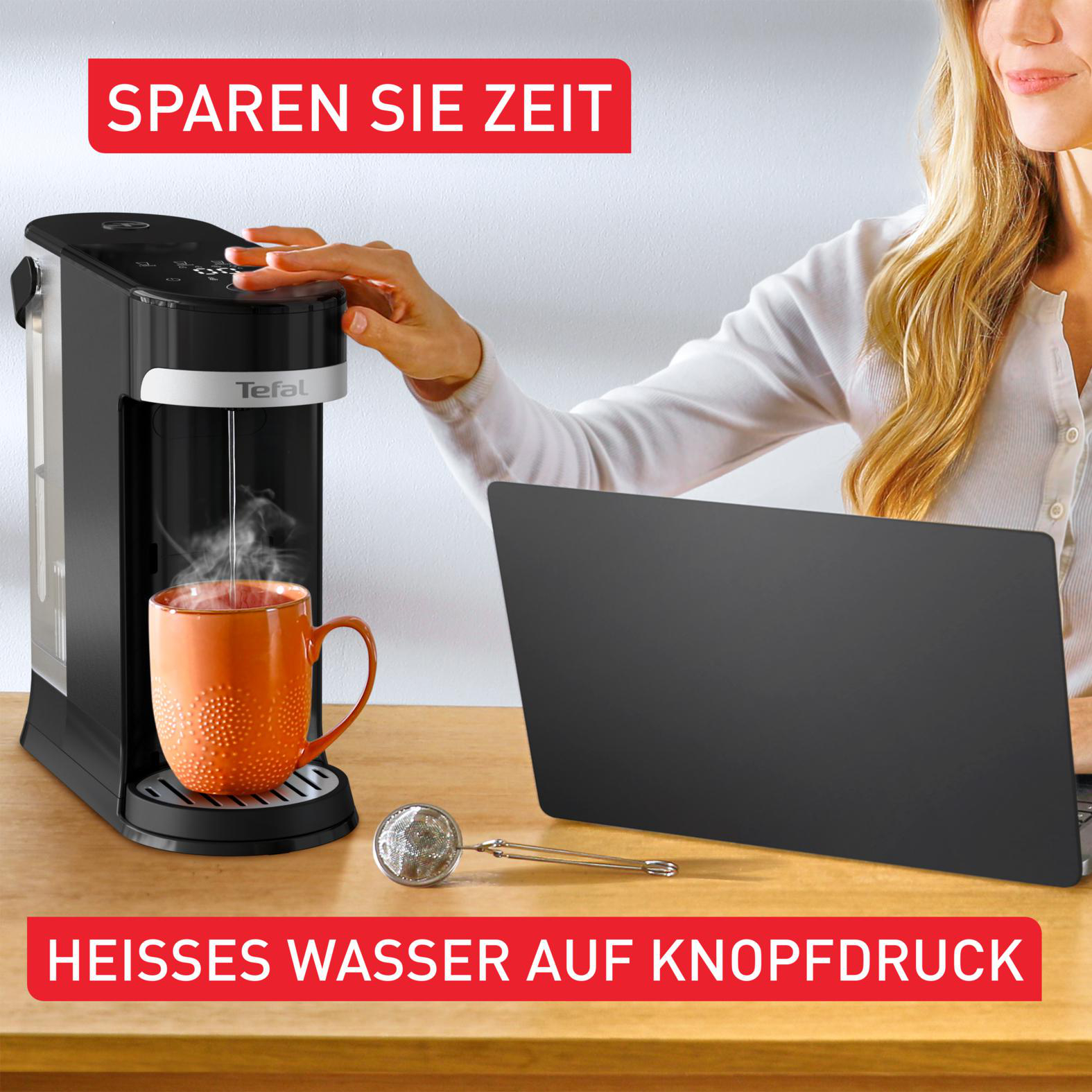 Eine Frau bedient eine schwarze Kaffeemaschine und füllt eine orangefarbene Tasse. Auf dem Holztisch liegen ein Laptop und ein Tee-Ei.