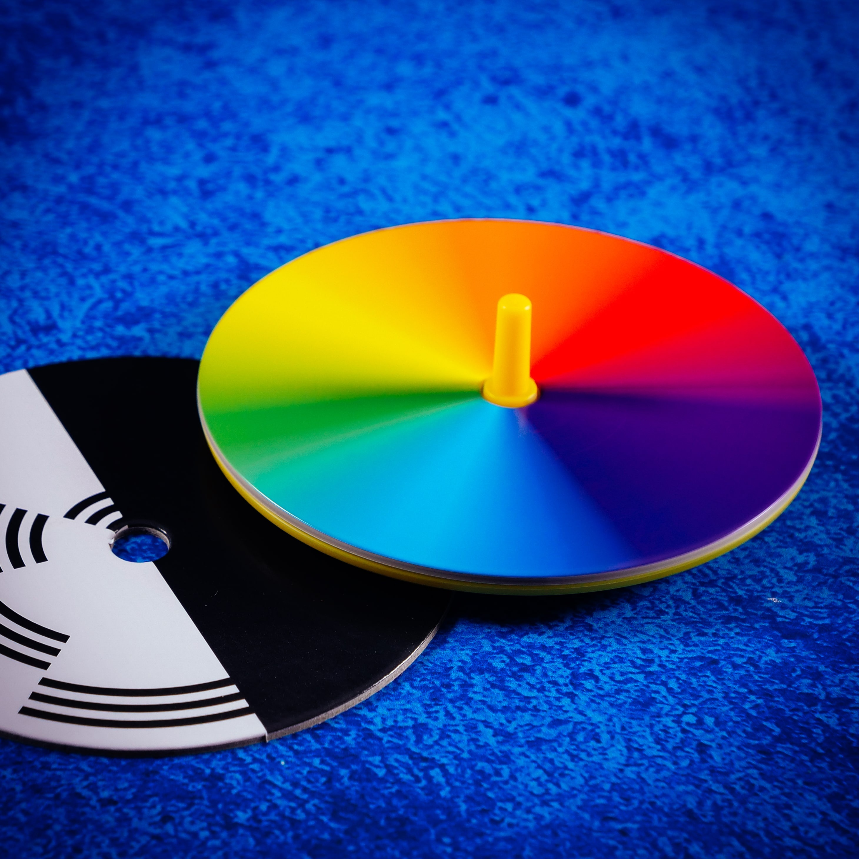 Kolorowy dysk z żółtym kołkiem i czarno-białym dyskiem na niebieskim, teksturowanym tle.