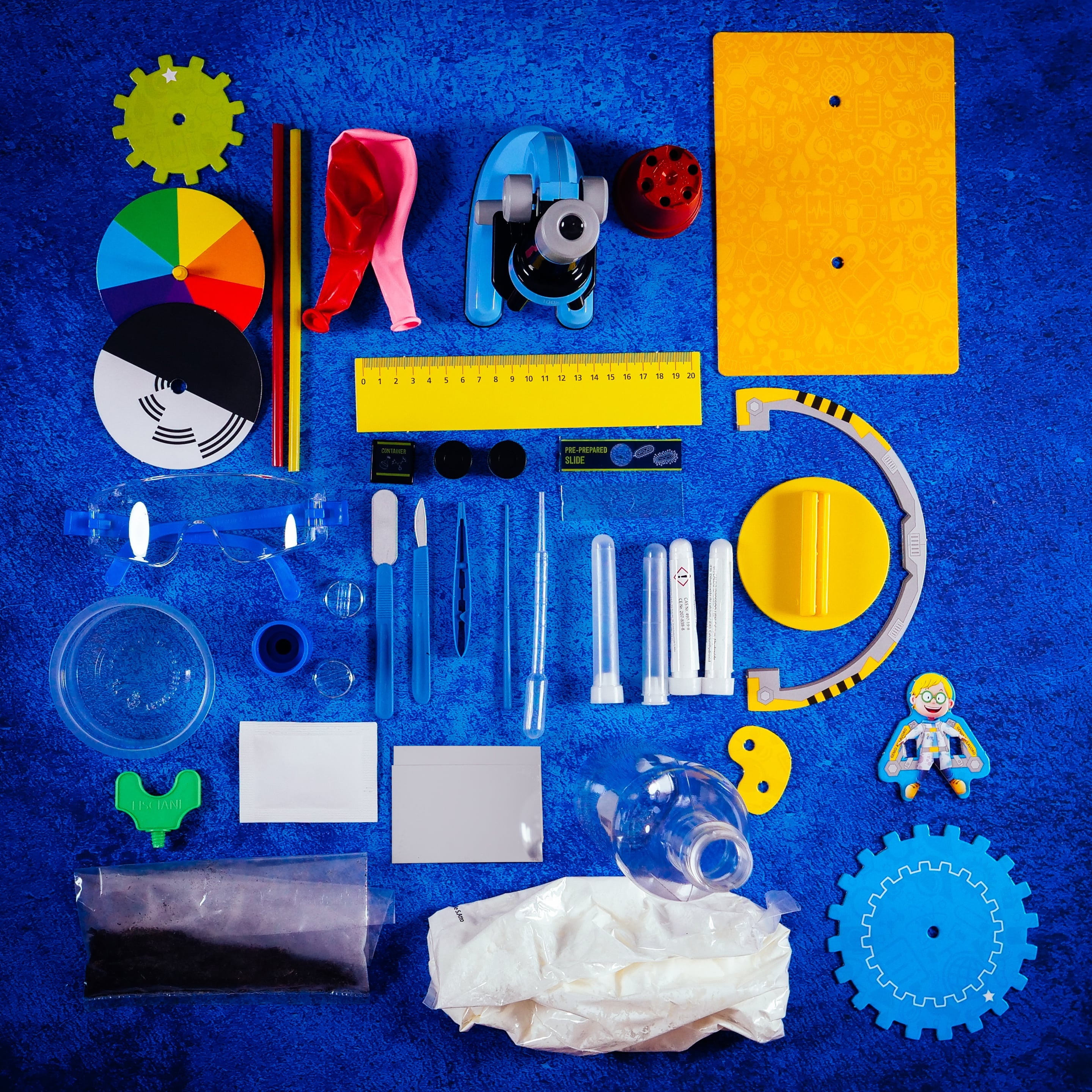 Różne elementy zestawu naukowego na niebieskiej powierzchni: koła zębate, linijka, probówki, balon i sprzęt laboratoryjny.