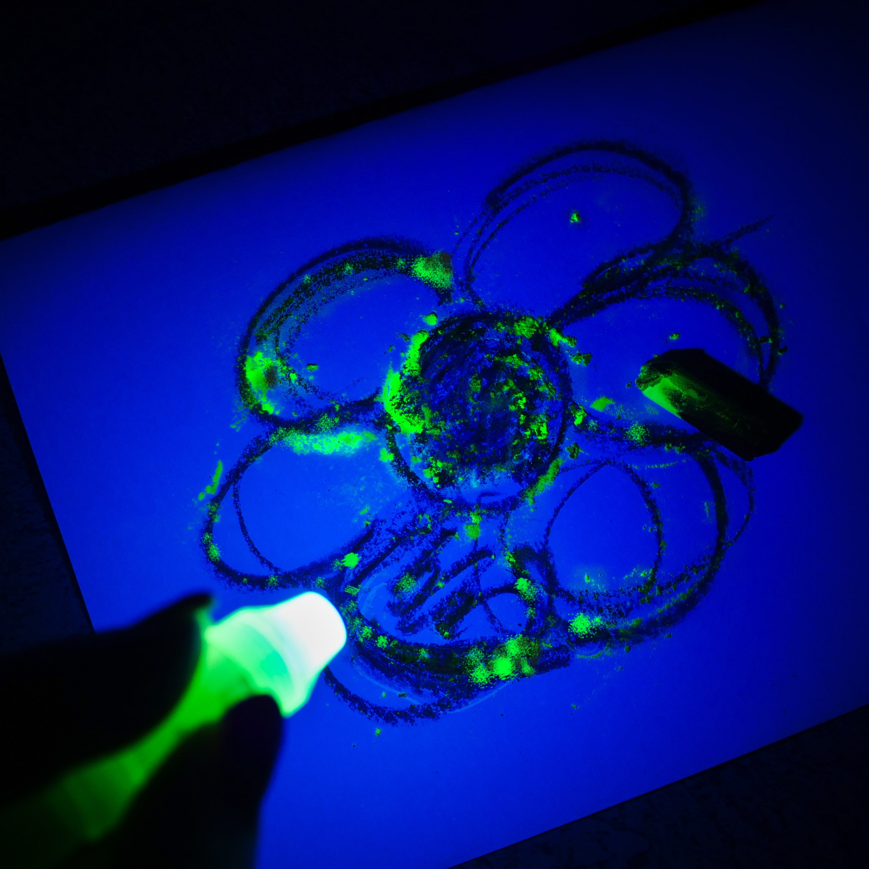 Świecący rysunek kwiatu na niebieskim papierze. Ręka trzyma zielone światło. Widoczna jest czarna gumka. Scena jest ciemna.