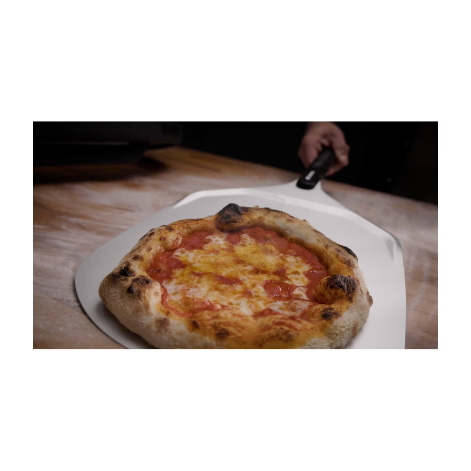 Una pizza con salsa al pomodoro e formaggio su una pala per pizza. Una mano tiene la pala. La pizza è su legno.