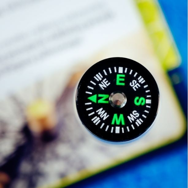 Czarny kompas z zielonymi literami leży na książce. Igła kompasu wskazuje północ. Tło to niebieska powierzchnia.