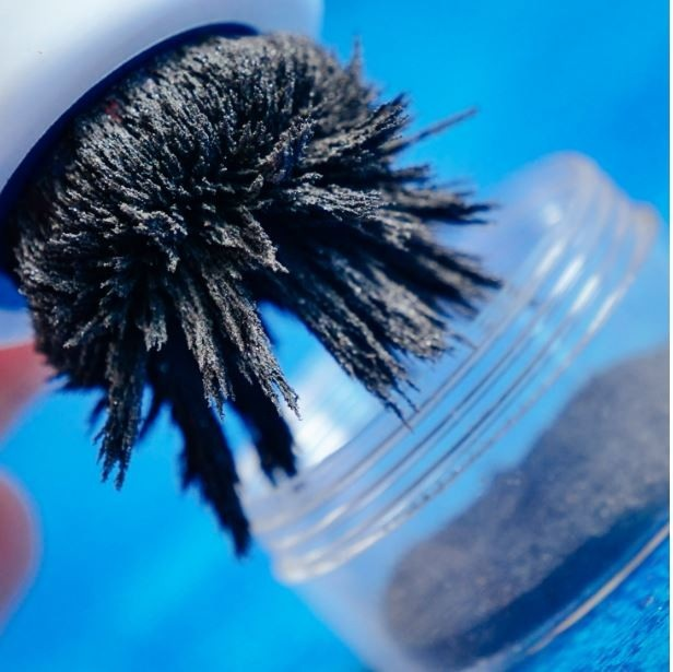 Ręka trzyma słoik z masą czarnego proszku, prawdopodobnie opiłkami żelaza, przyciąganymi przez magnes. Niebieskie tło.