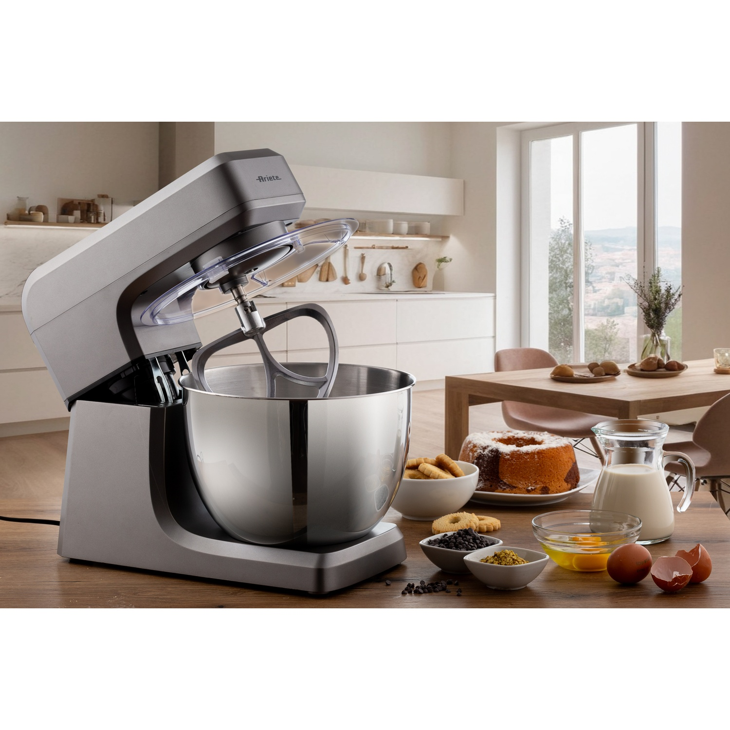 Scena di cucina con impastatrice, torta, latte, uova e ingredienti su un tavolo di legno. Una finestra offre una vista.