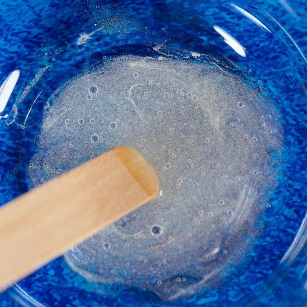 Zbliżenie niebieskiej miski z przezroczystą, pieniącą się substancją. Jasnobrązowy drewniany patyczek miesza zawartość.
