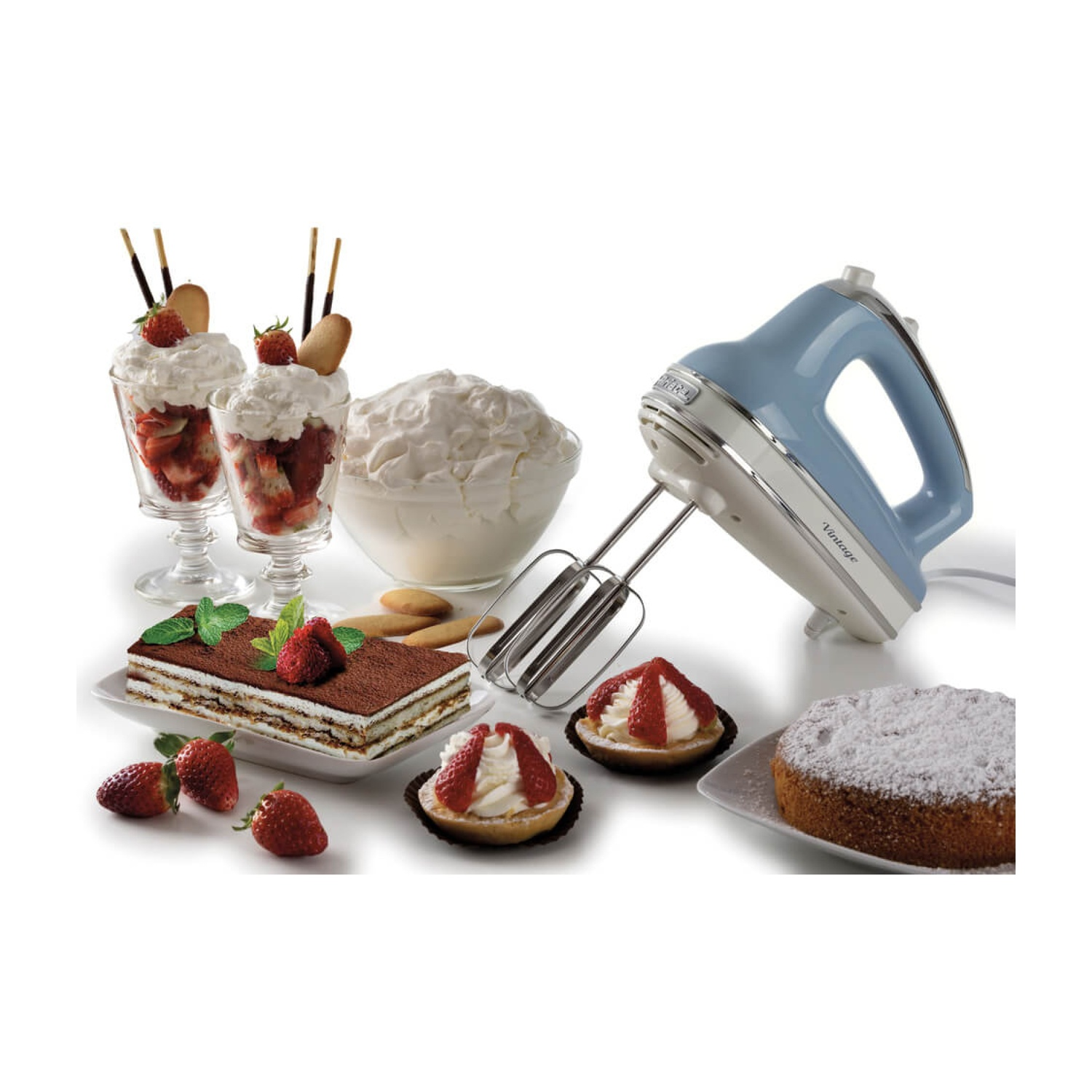 Un frullatore a mano azzurro chiaro è appoggiato vicino a dessert, tra cui parfait alla fragola, tiramisù e crostate, su una superficie bianca.