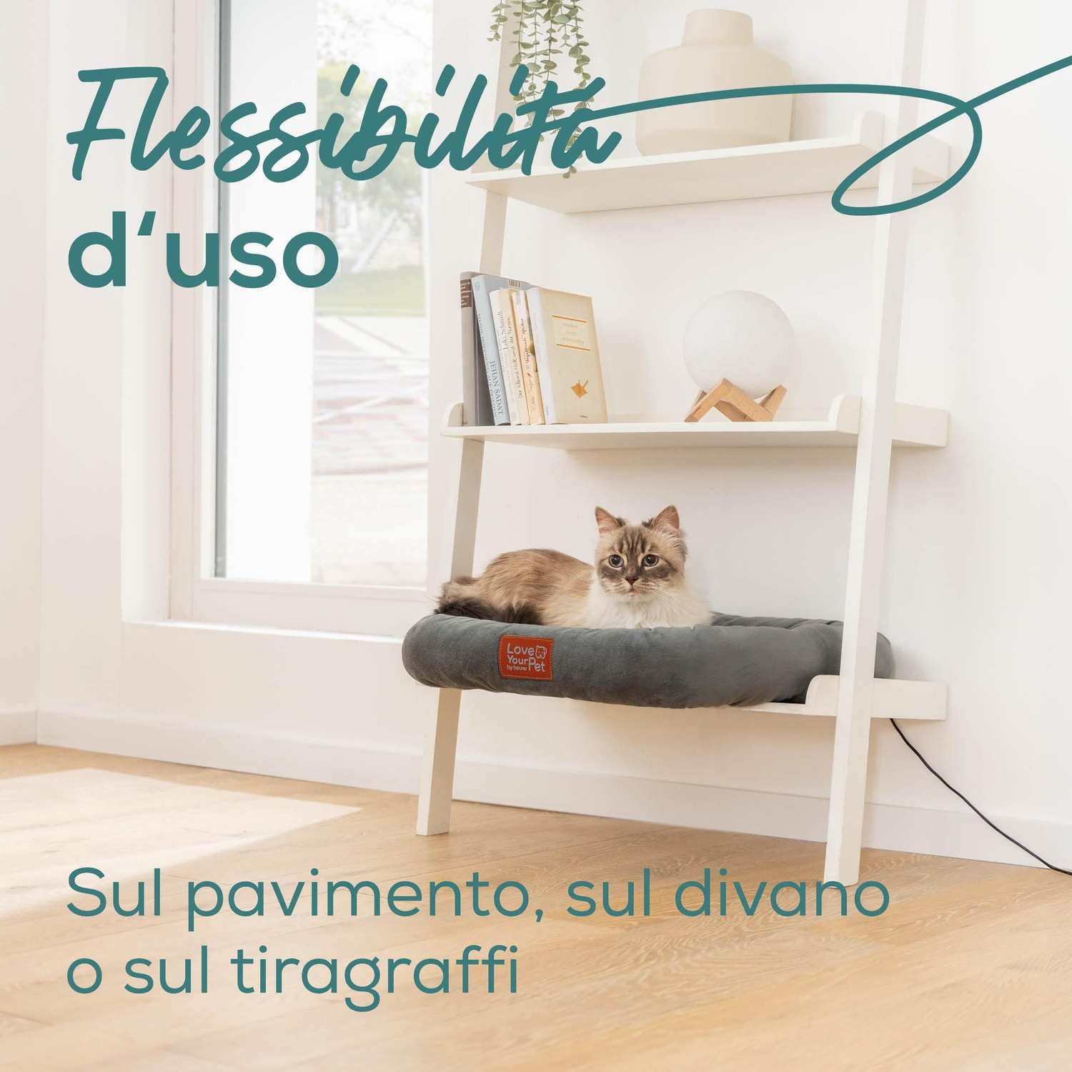 Un gatto riposa su un letto grigio su una mensola bianca. La mensola ha libri e una lampada. Il testo dice: Flessibilita d'uso, Sul pavimento, sul divano o sul tiragraffi.