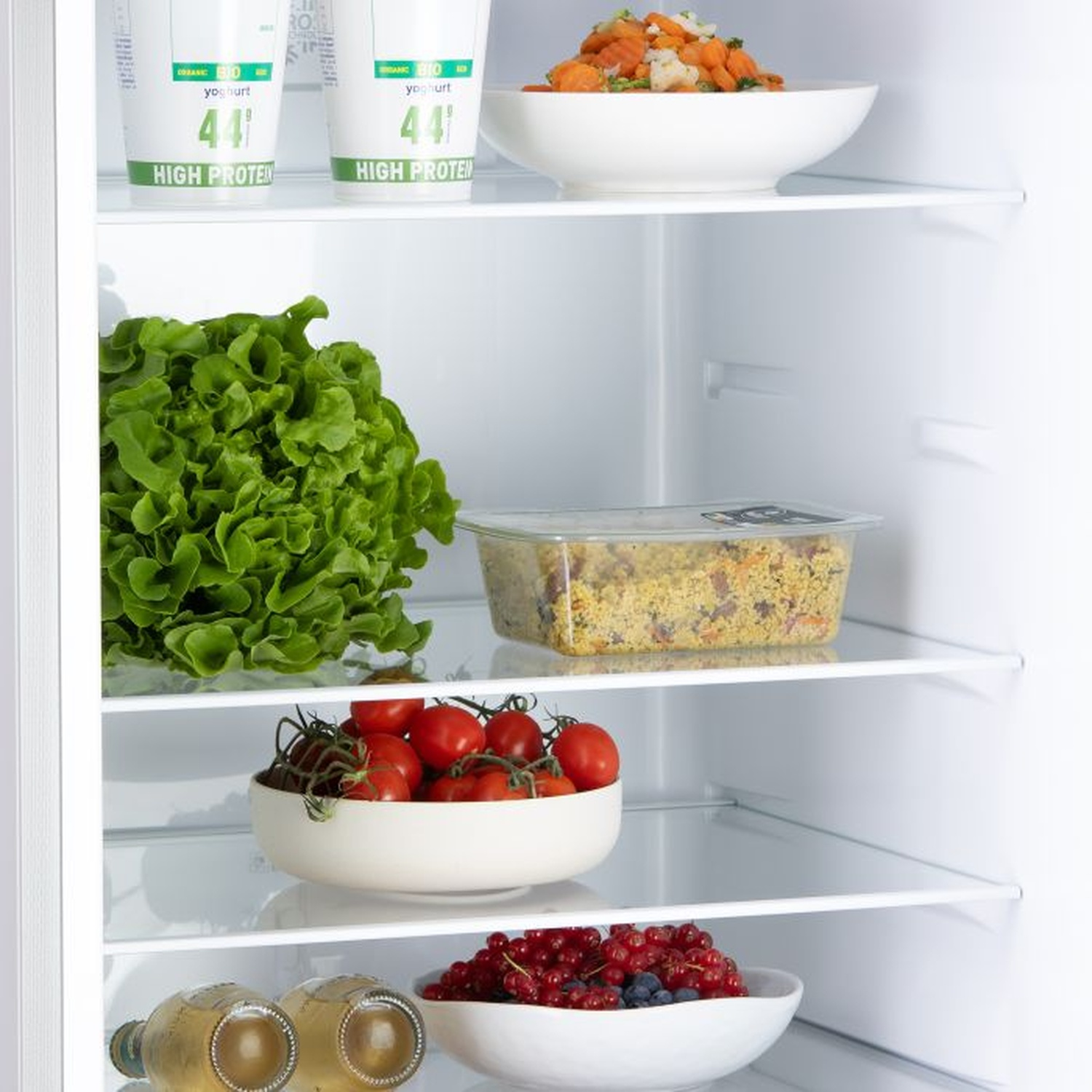 À l'intérieur d'un réfrigérateur: yaourt, salade, tomates, légumes cuits, baies et bouteilles. Divers aliments sont stockés dans des bols et des contenants blancs.