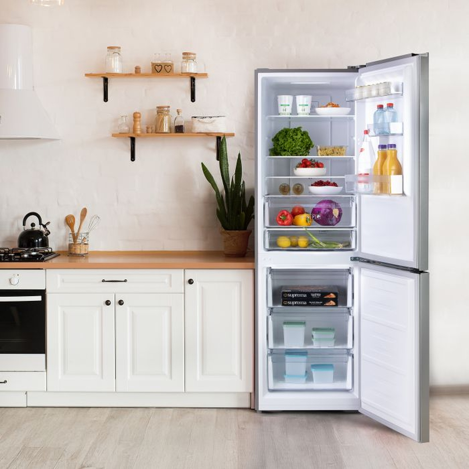Réfrigérateur ouvert rempli de nourriture. La cuisine comprend une cuisinière, des armoires, des étagères avec des bocaux et une plante. Le sol est en bois clair.