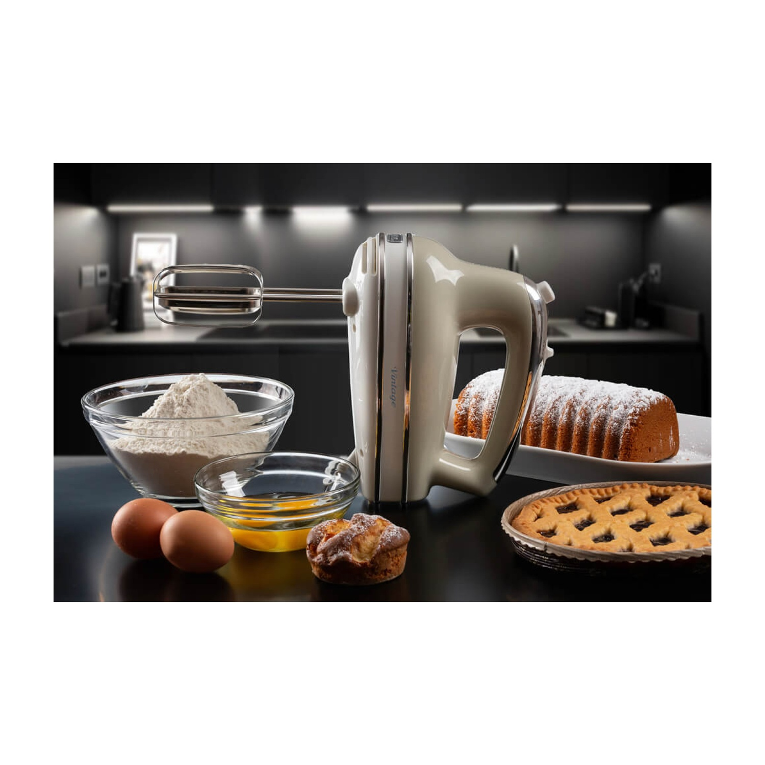 Un mixer color crema su un bancone con ingredienti: farina, uova e una torta. Sono presenti anche una torta e un muffin.