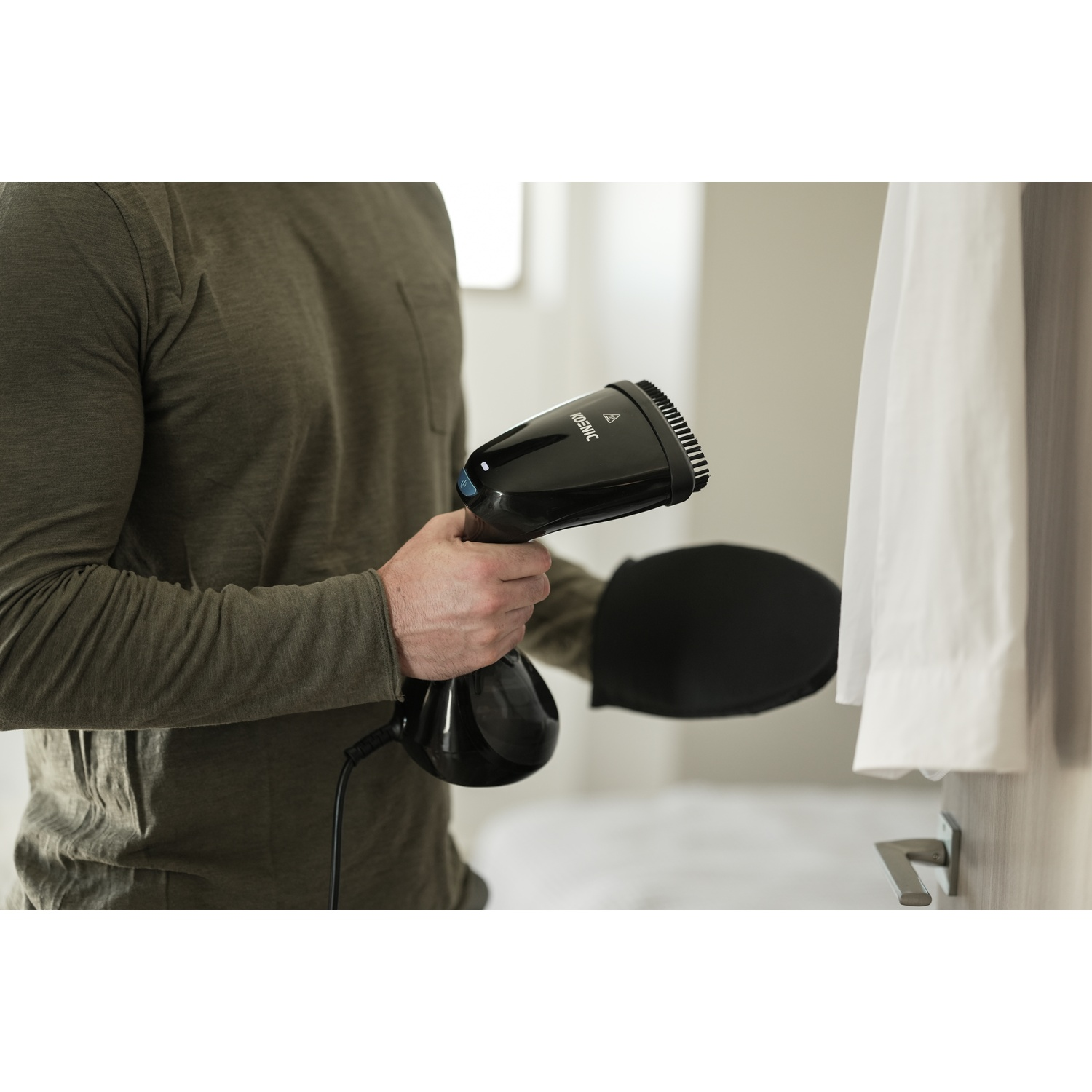 Una persona sta stirando una camicia bianca con un vaporizzatore nero. La persona indossa una camicia verde. Un cuscinetto nero è accanto alla camicia.