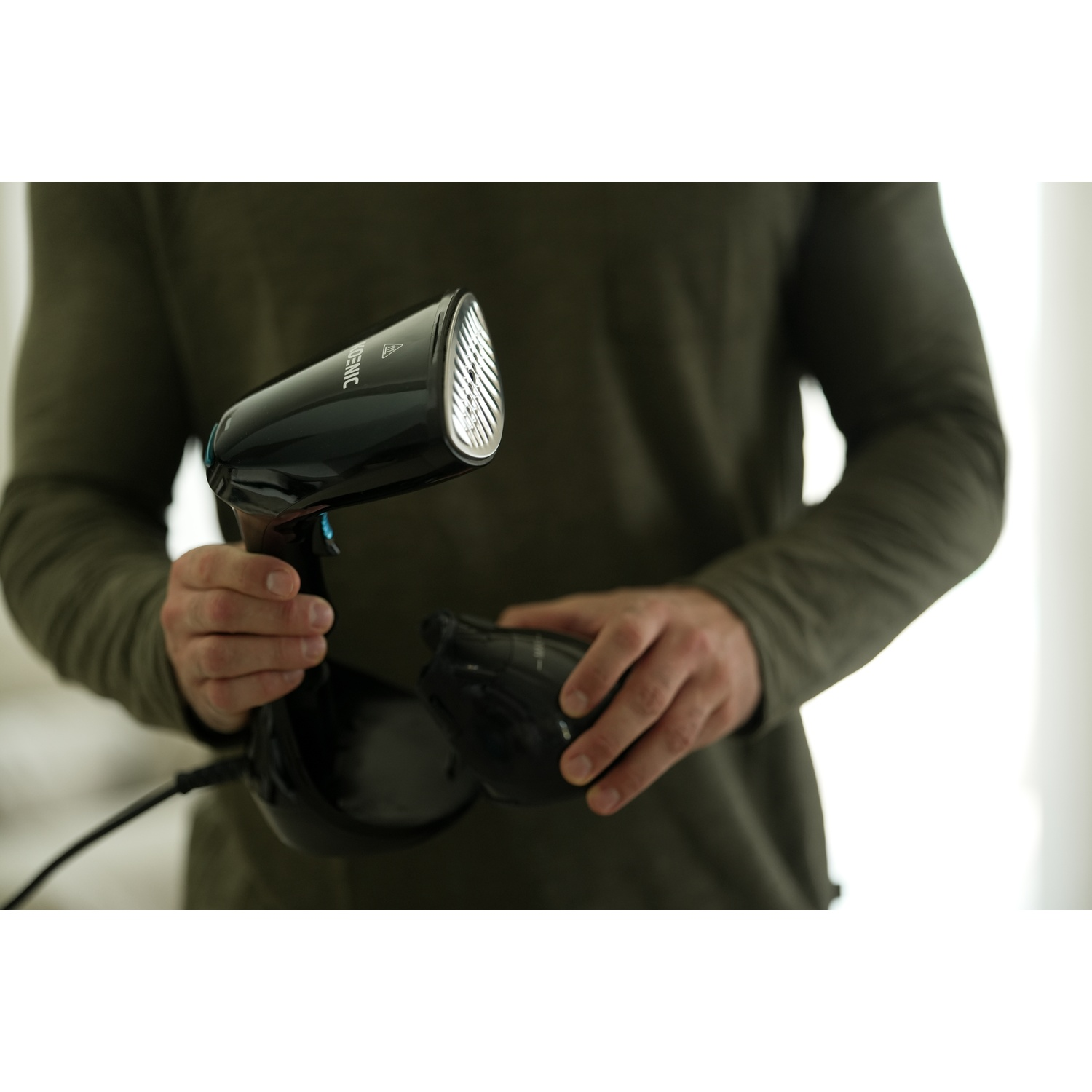 Una persona tiene in mano uno steamer nero per indumenti. Lo steamer ha un ugello e un'impugnatura. Lo sfondo è sfocato.