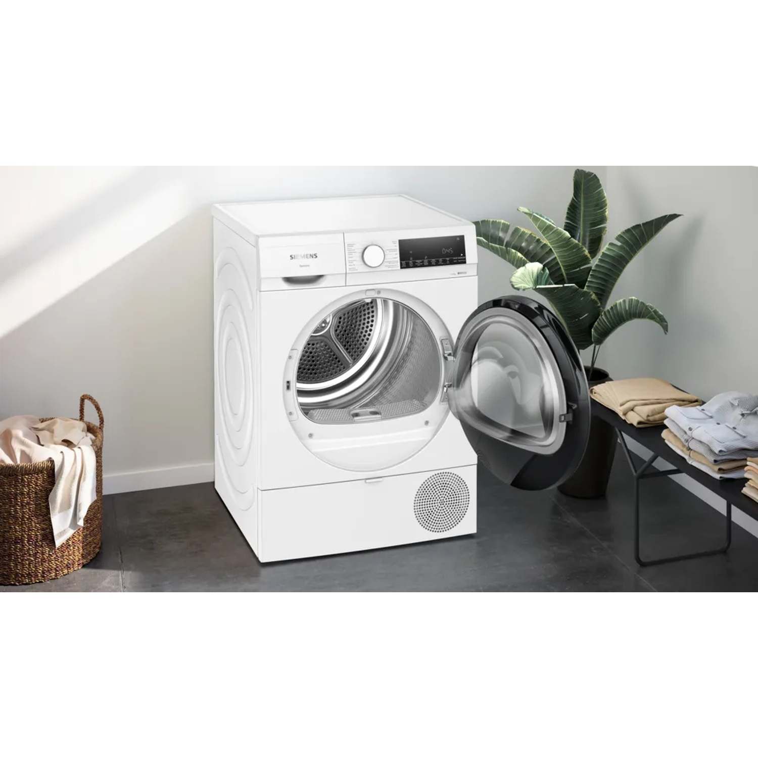A white dryer with the door open. There is a basket with clothes to the left. A plant and folded clothes are to the right.