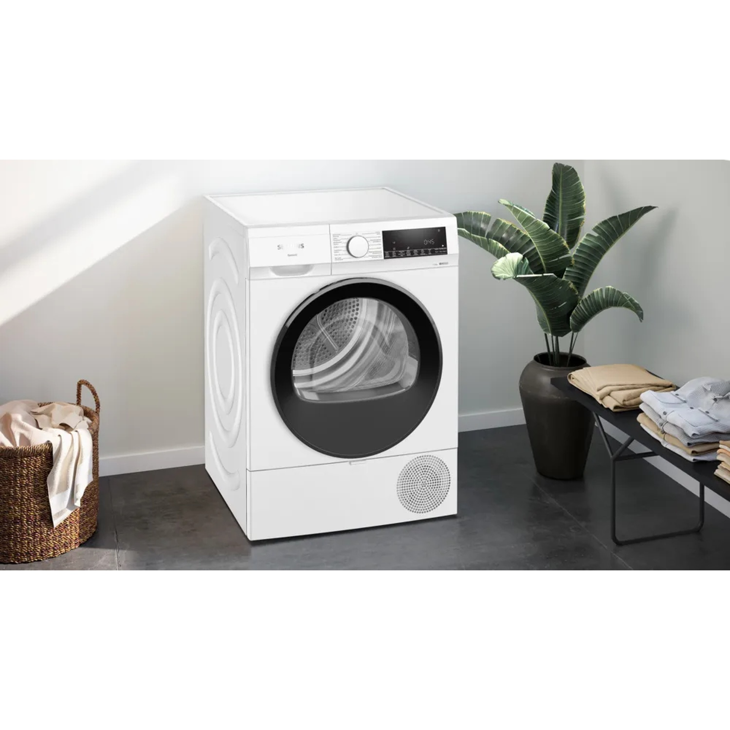 A white dryer with a black door, next to a laundry basket and a plant. Clothes are folded on a black table.