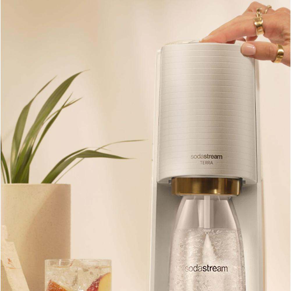 Egy kéz megnyom egy gombot egy fehér SodaStream gépen. Előtérben egy pohár gyümölcsös víz, balra növények.