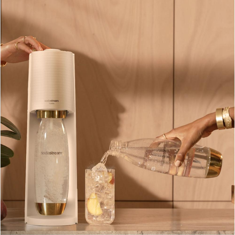 Egy SodaStream gép egy pohár jéggel és gyümölccsel van. Egy kéz vizet önt egy üvegből a pohárba.